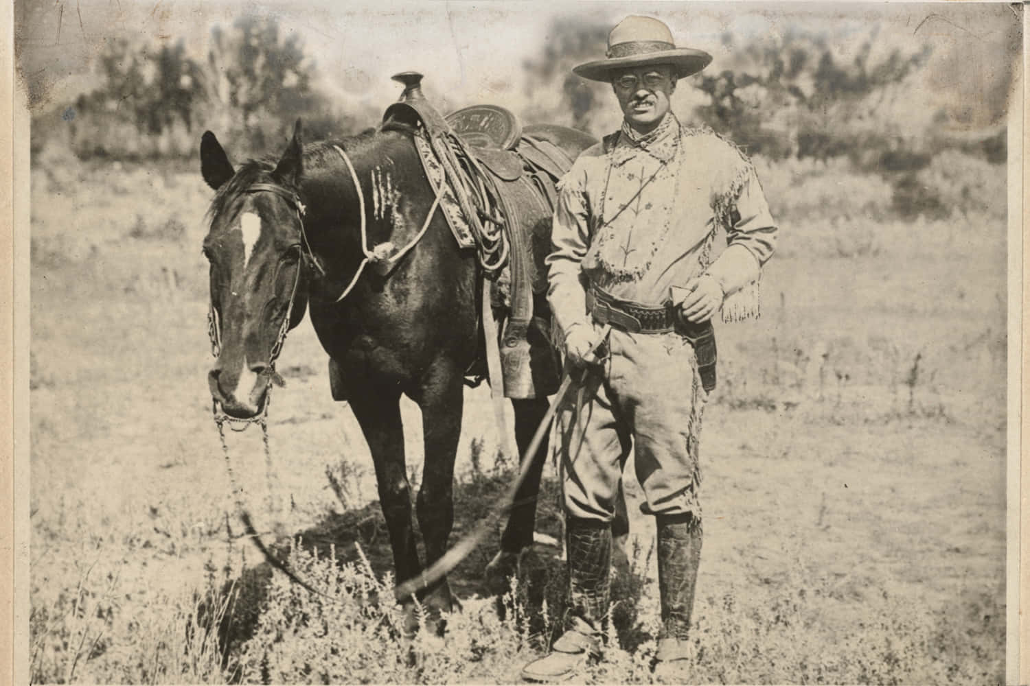 Old Western 桌布 壁紙 — 一張復古棕褐色調的照片，一名穿著牛仔服飾的男子站在一匹備好鞍的馬旁邊，背景是乾燥的草地。