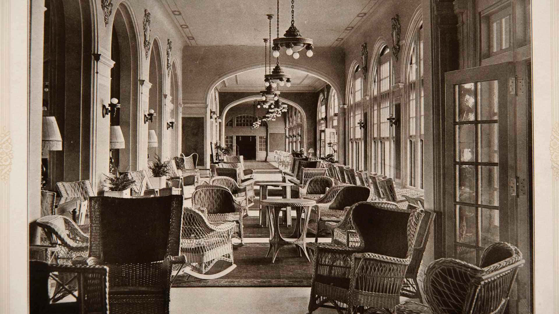 Haunted Hotels 桌布 壁紙 — 一張復古棕褐色調的照片，展示了一條優雅的長走廊，兩旁擺放著藤椅和桌子，上方是拱形窗戶和吊燈。