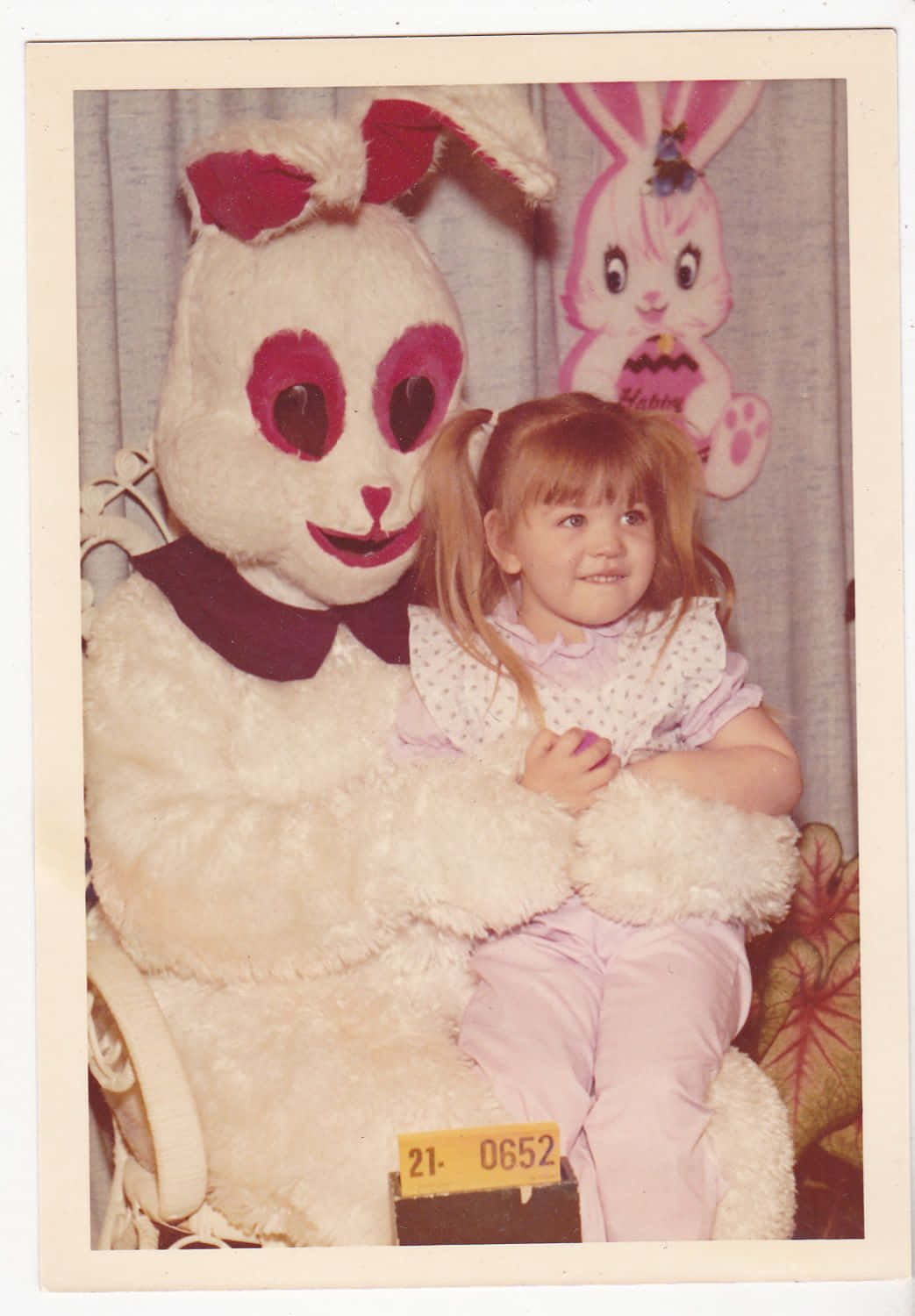 Creepy Easter Bunny 桌布 壁紙 — 一張復古照片，一個穿著粉紅色衣服的微笑小孩坐在一個帶有紅色蝴蝶結和心形眼睛的白色大型復活節兔子服裝上。