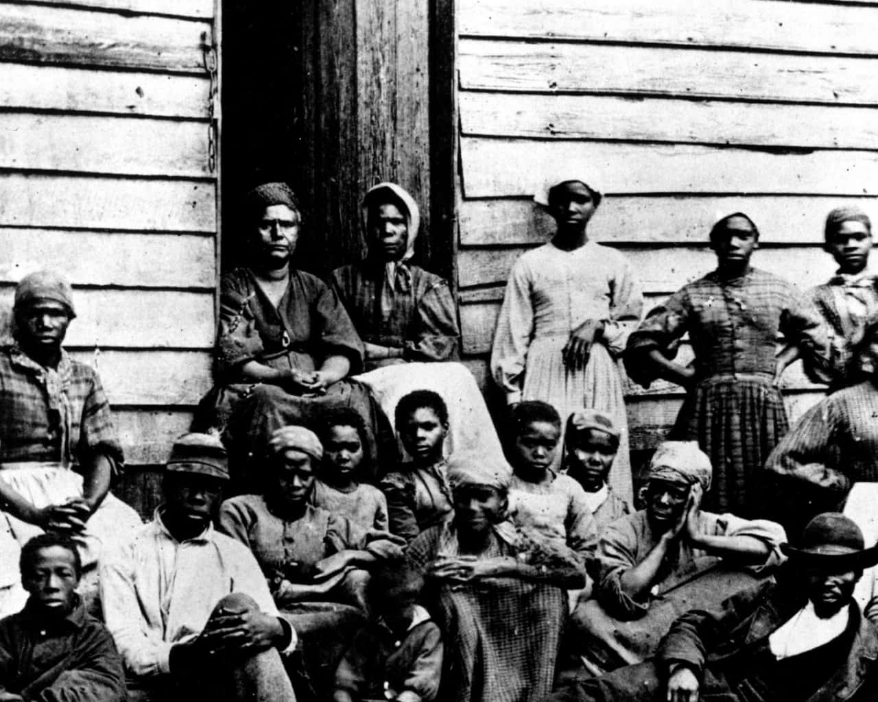Juneteenth 桌布 壁紙 — 一張復古黑白照片，一大群非裔美國成年人和兒童在一扇風化的木屋門前莊嚴地擺姿勢。