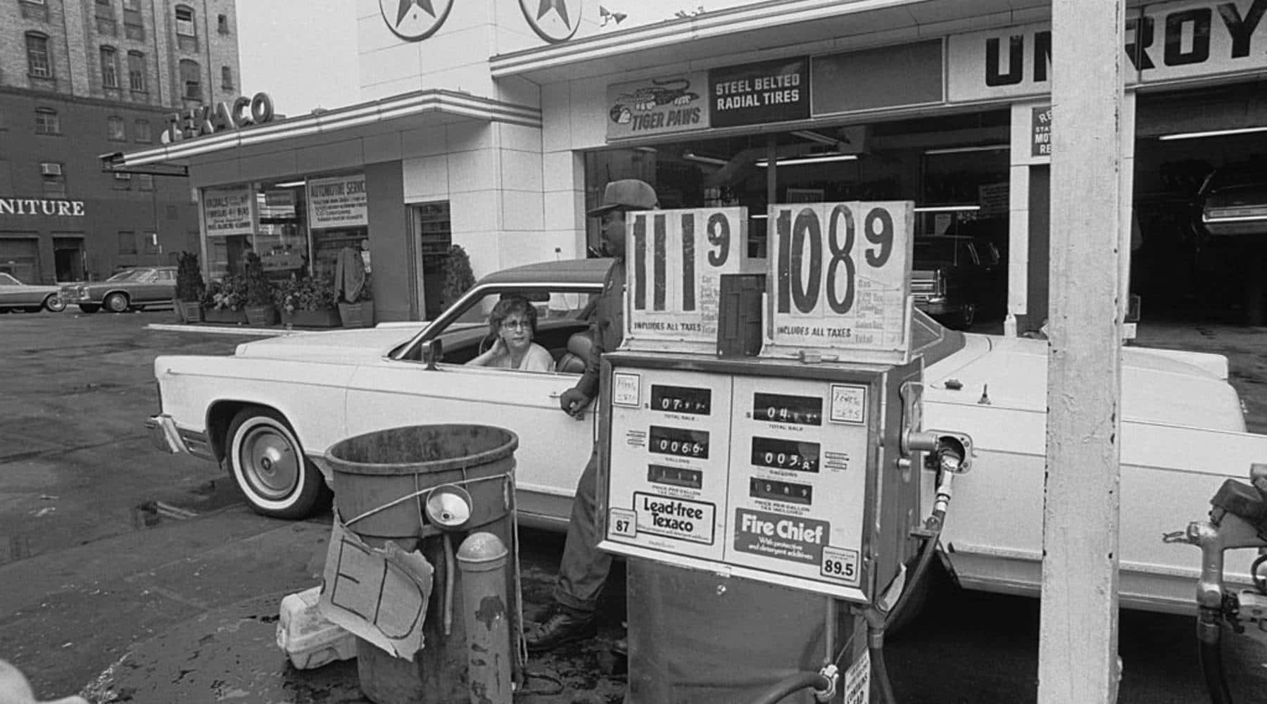 1970s 桌布 壁紙 — 一張復古黑白照片，顯示一位女士在復古加油站油泵旁，旁邊有一輛經典汽車和價格標牌。