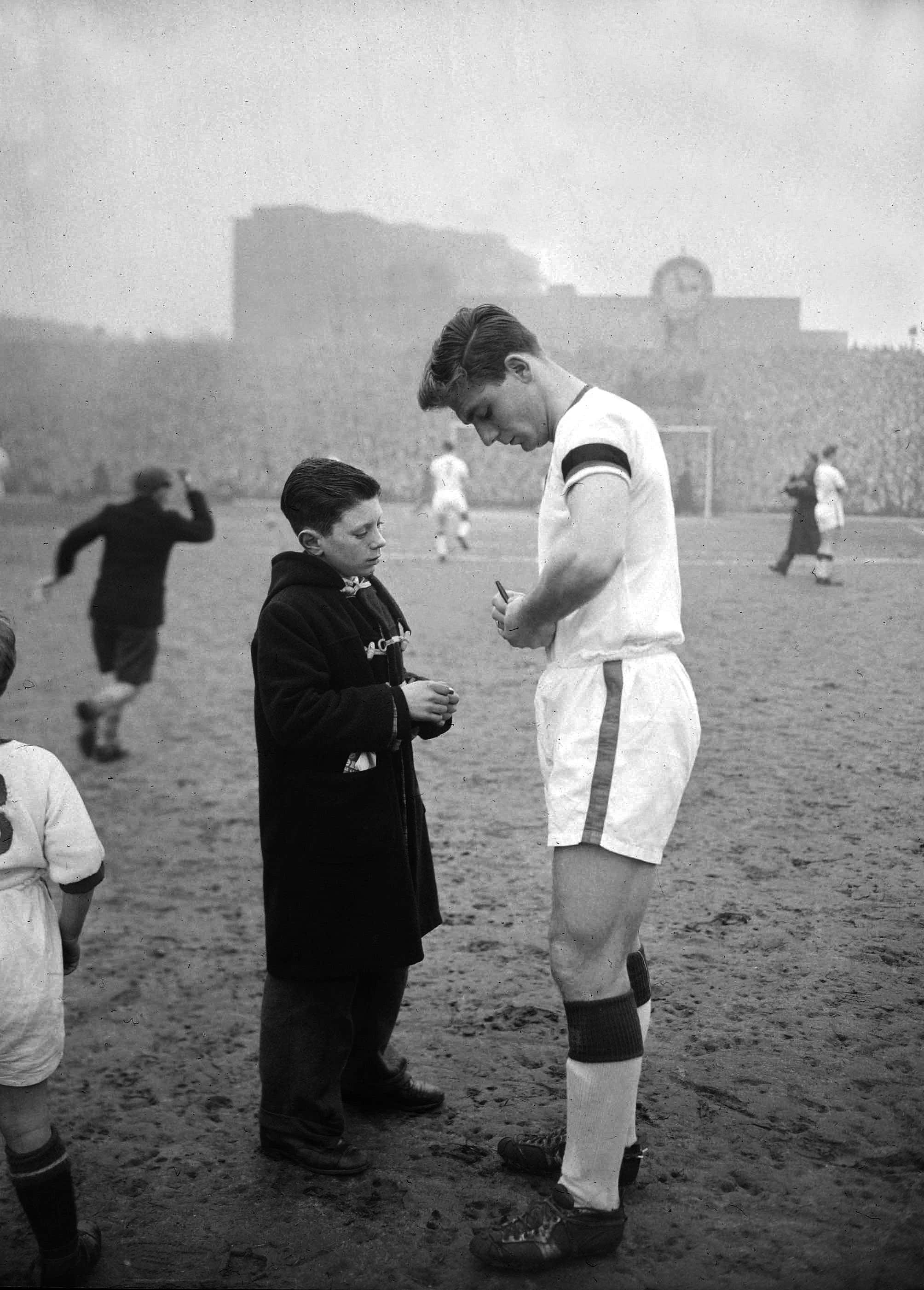 Football Players 桌布 壁紙 — 一張復古黑白照片顯示，一名穿著外套的小男孩在泥濘的球場上與一名高大的足球運動員面對面站立，當時正在進行比賽。