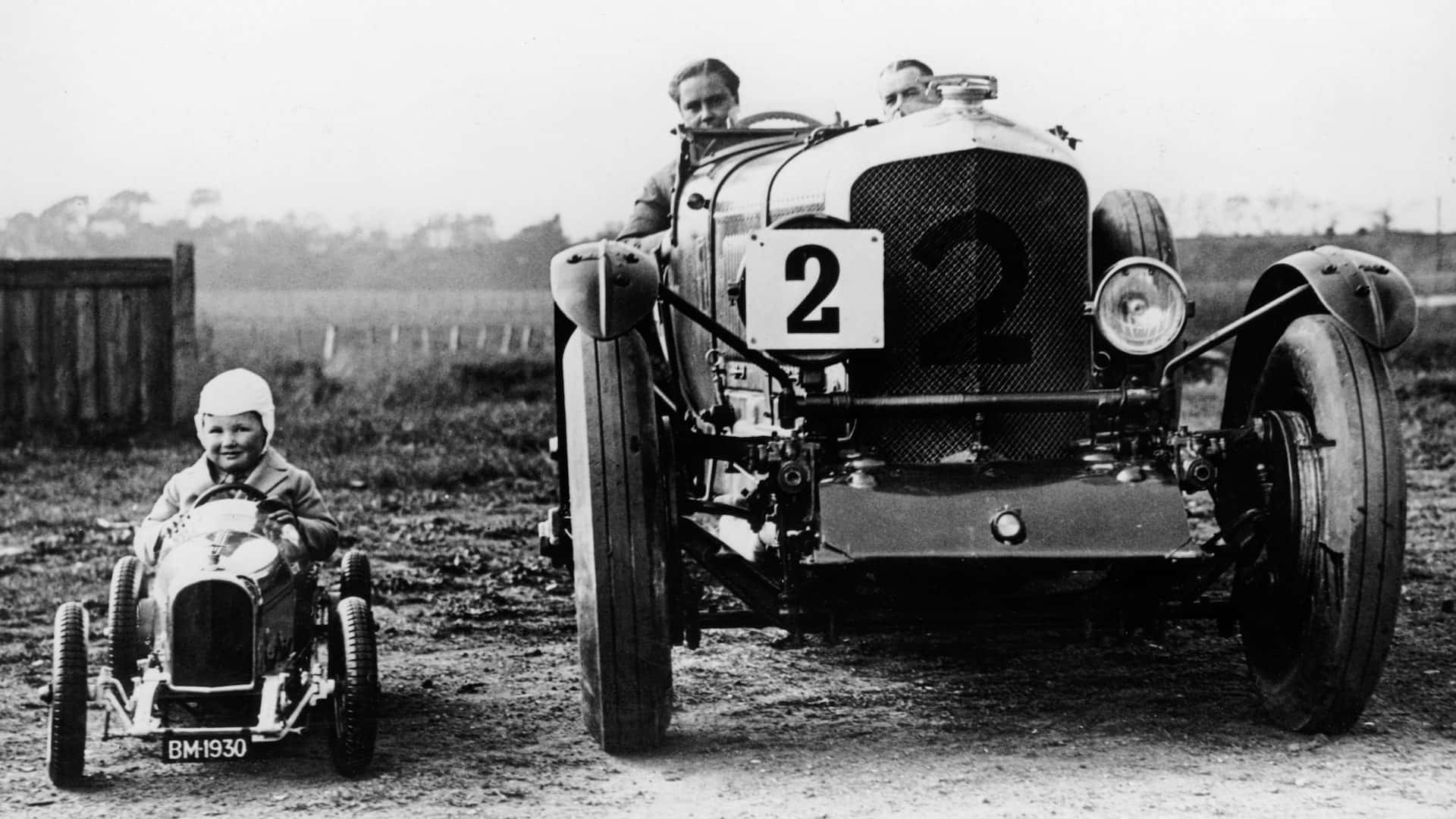 Black And White Vintage Car 桌布 壁紙 — 一張復古黑白照片顯示一個微笑的孩子坐在微型賽車旁，旁邊是一輛帶有2號的大型復古賽車。