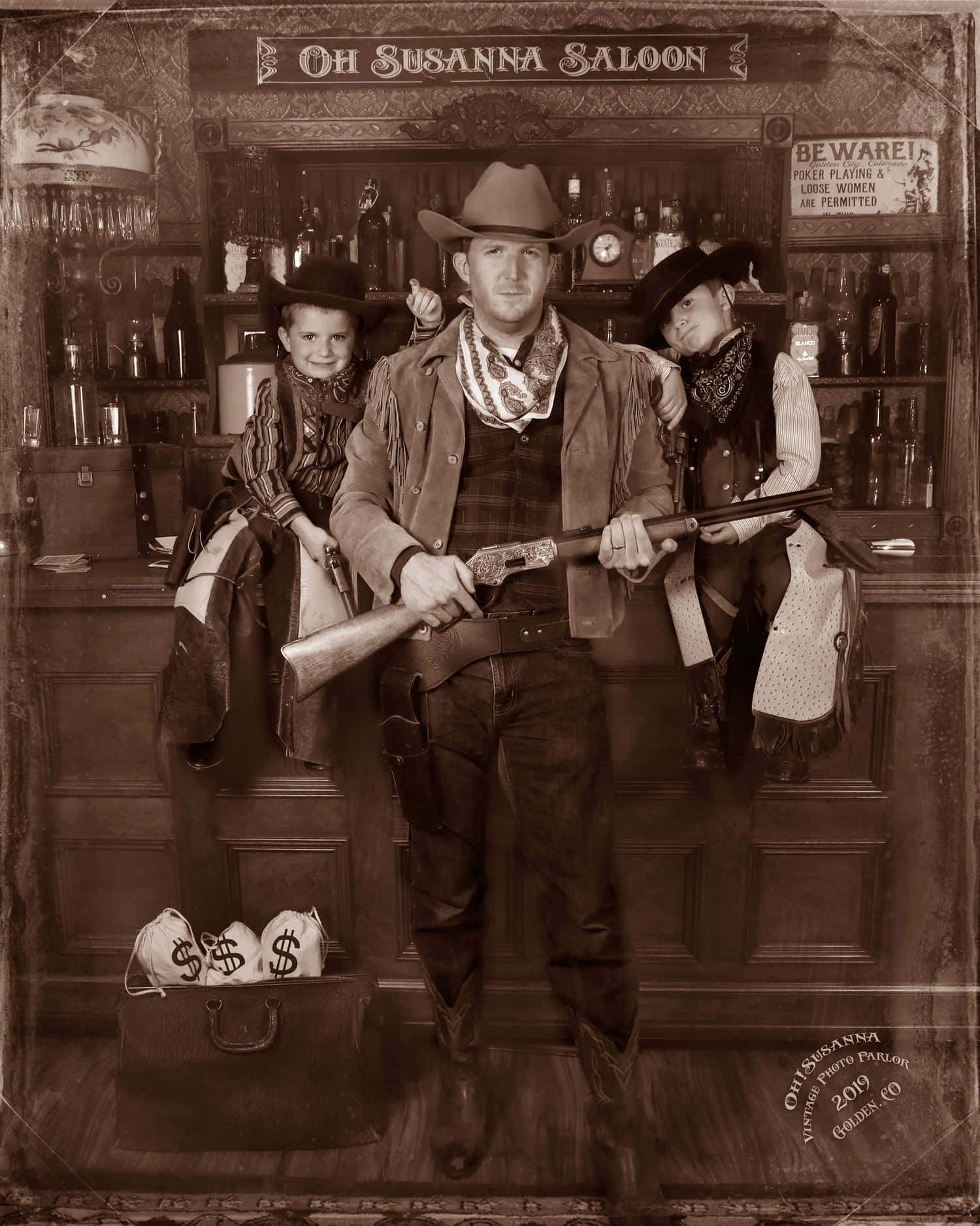 Old Western 桌布 壁紙 — 一張懷舊色調的照片，一名牛仔與兩個孩子在復古沙龍中擺姿勢，周圍有槍支和錢袋。