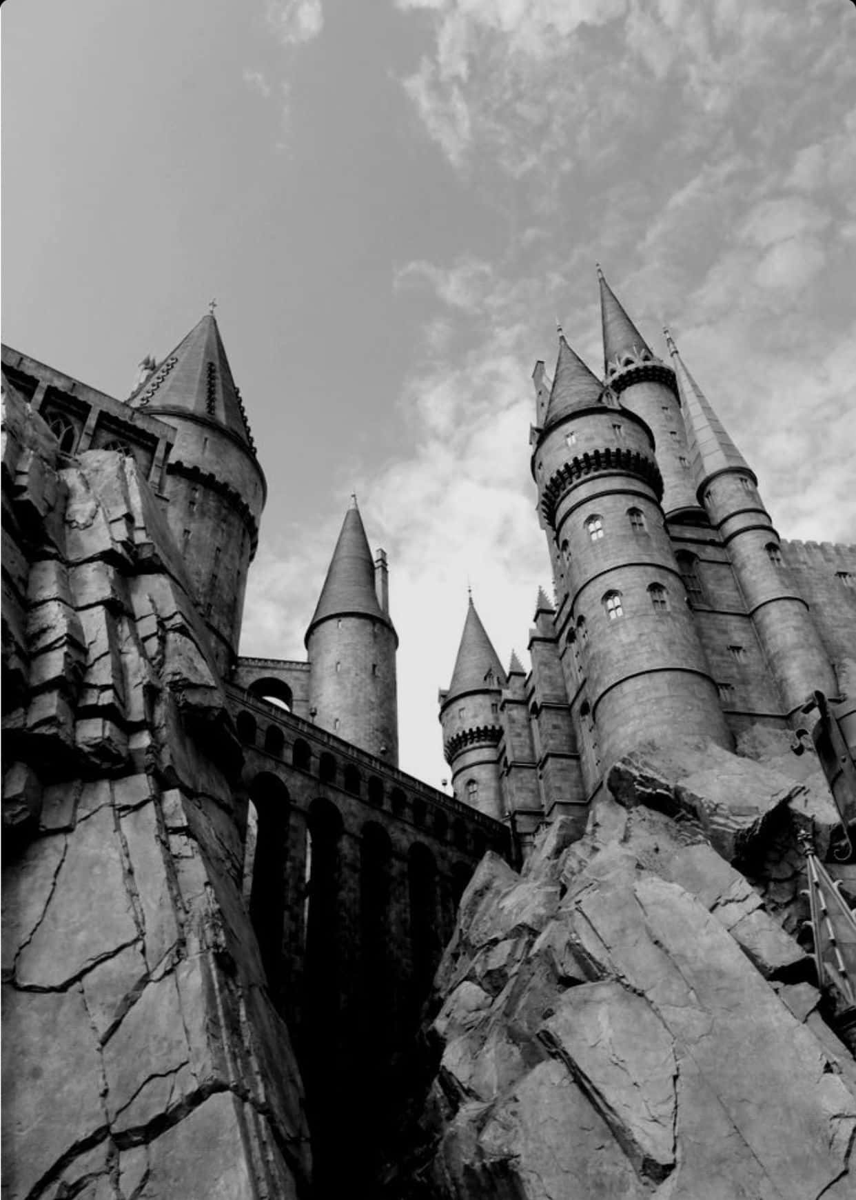 Harry Potter Black And White 桌布 壁紙 — 一張戲劇性的低角度黑白照片，展示霍格華茲城堡坐落在崎嶇懸崖上，天空多雲。
