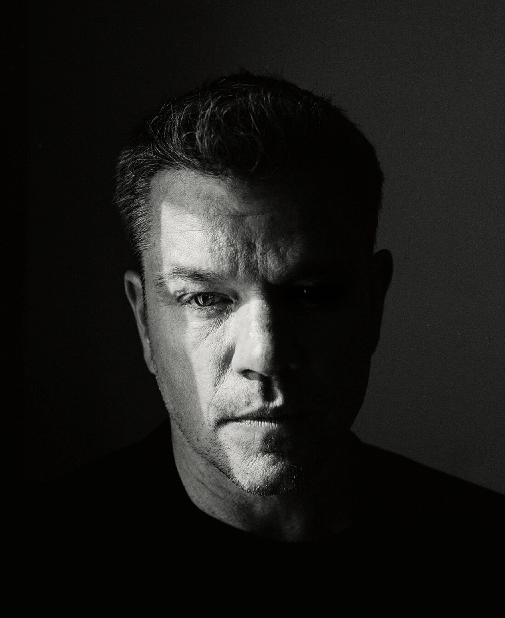 Matt Damon 桌布 壁紙 — 一張戲劇性的黑白人像照，捕捉一名眼神銳利、帶有鬍渣的男子，在強烈側光照射下，背景為深色。