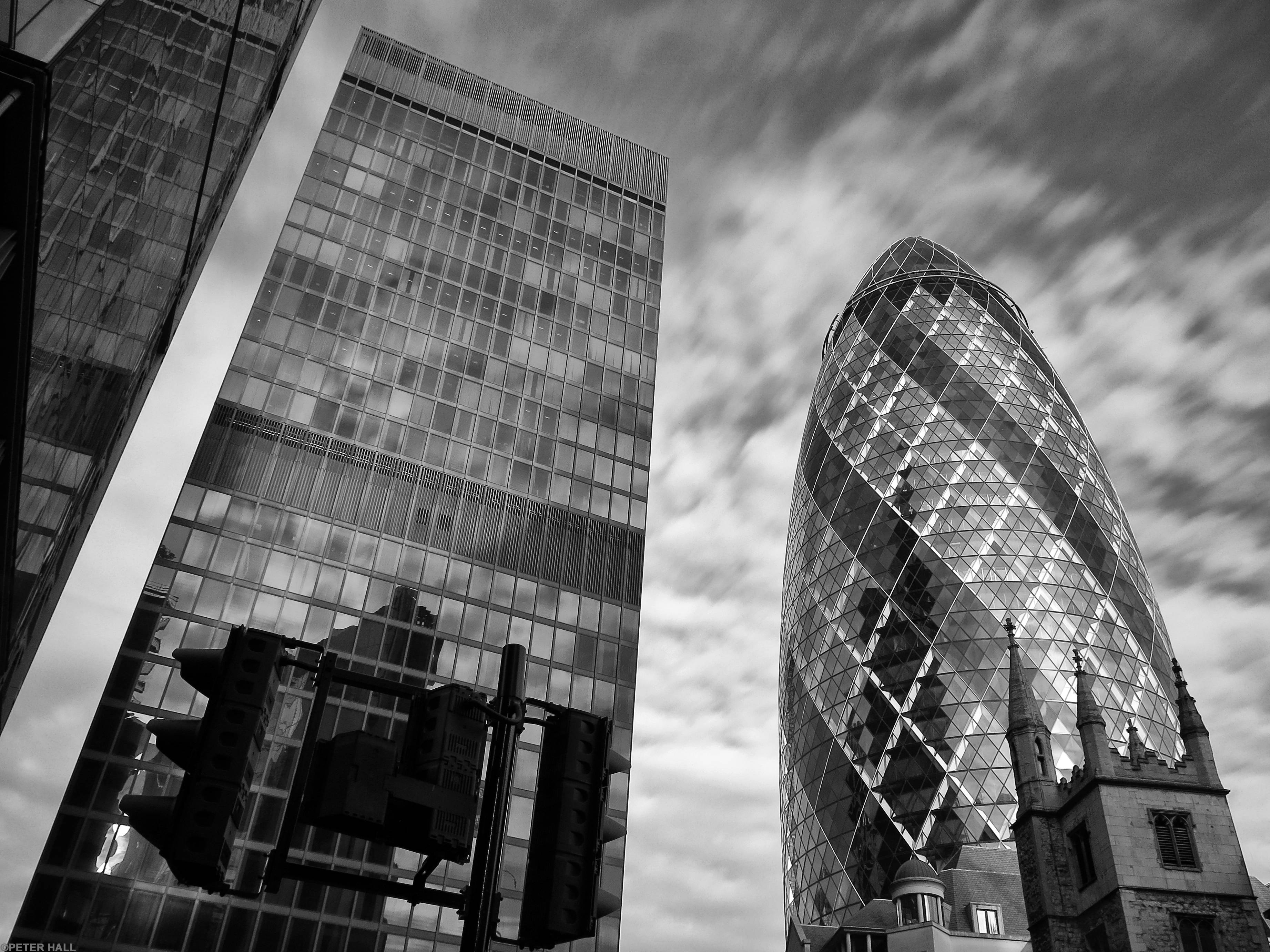 Buildings 桌布 壁紙 — 一張戲劇性的黑白低角度照片，拍攝了倫敦標誌性的小黃瓜摩天大樓旁邊的一座傳統教堂，背景是陰天。