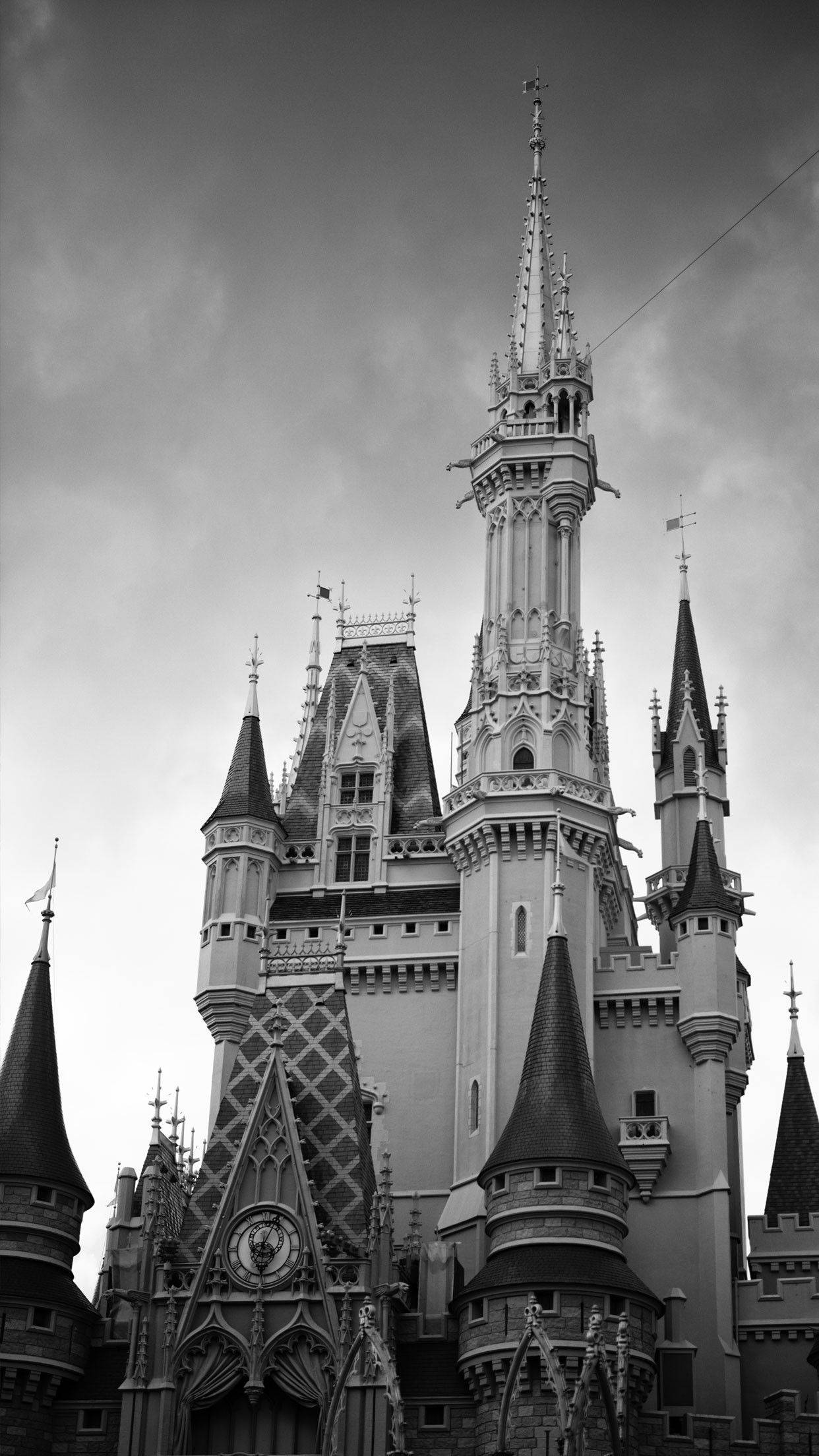 Disneyland Castle 桌布 壁紙 — 一張戲劇性的黑白低角度照片，拍攝了一座宏偉的童話城堡，尖塔聳立在多雲的天空下。