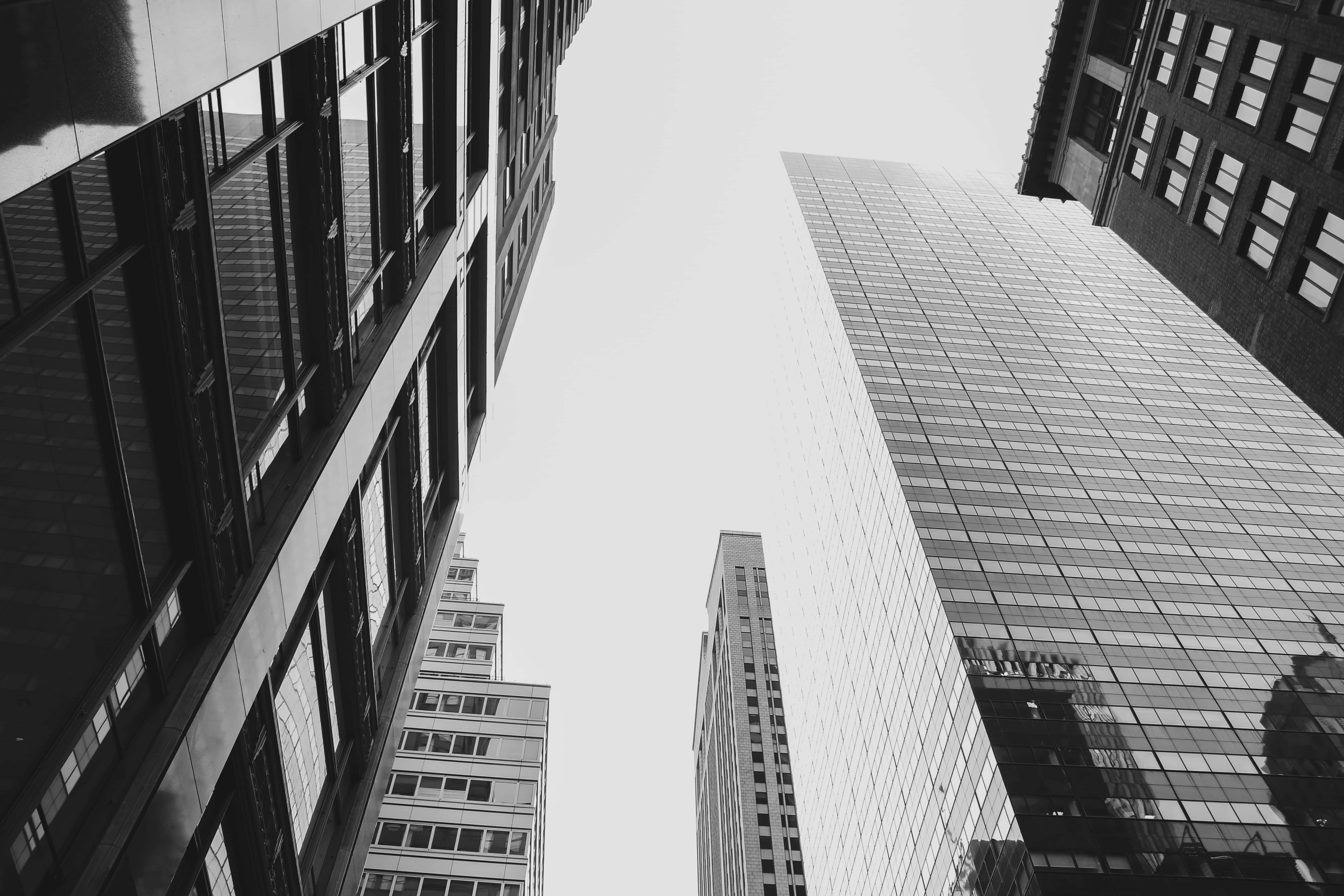 Buildings 桌布 壁紙 — 一張戲劇性的黑白低角度照片，拍攝了在密集的城市峽谷中，高聳的摩天大樓向明亮的天空匯聚。