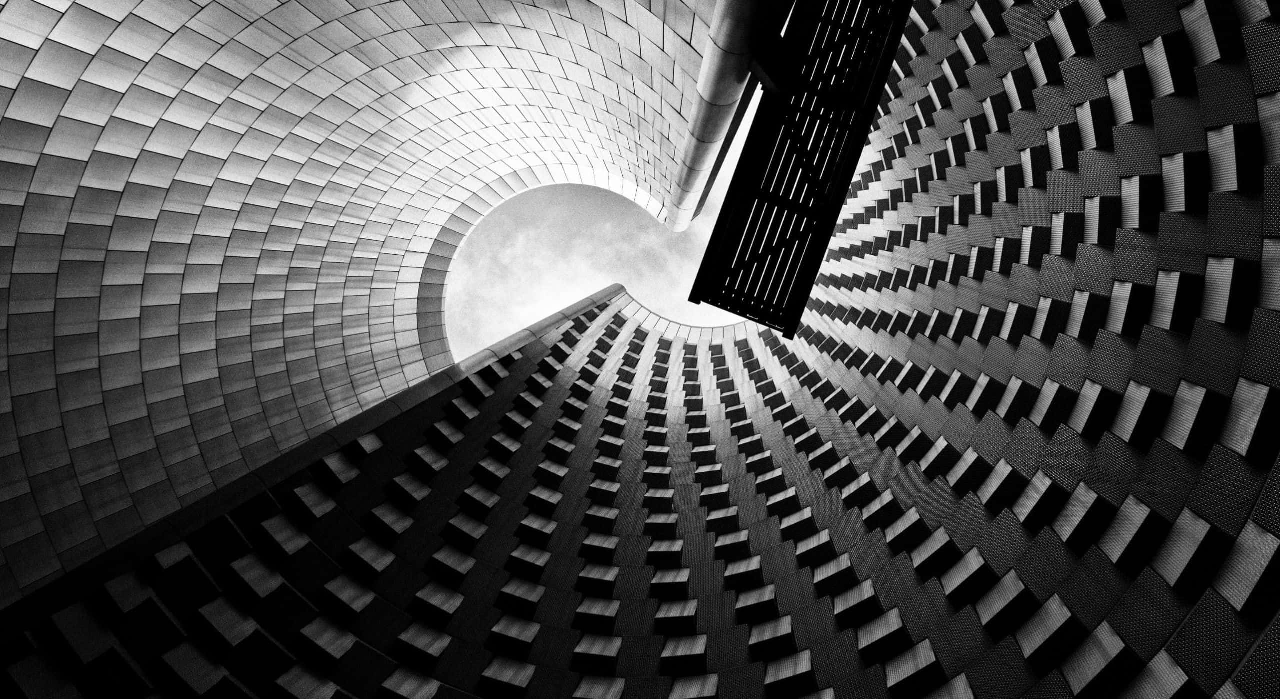 Black And White Architecture 桌布 壁紙 — 一張戲劇性的黑白低角度照片，捕捉了螺旋狀現代建築外牆向明亮圓形天空匯聚的畫面。