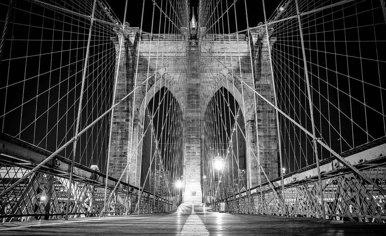 New York 桌布 壁紙 — 一張戲劇性的黑白夜景照片，仰望布魯克林大橋的石拱和鋼纜，朝向發光的燈光。