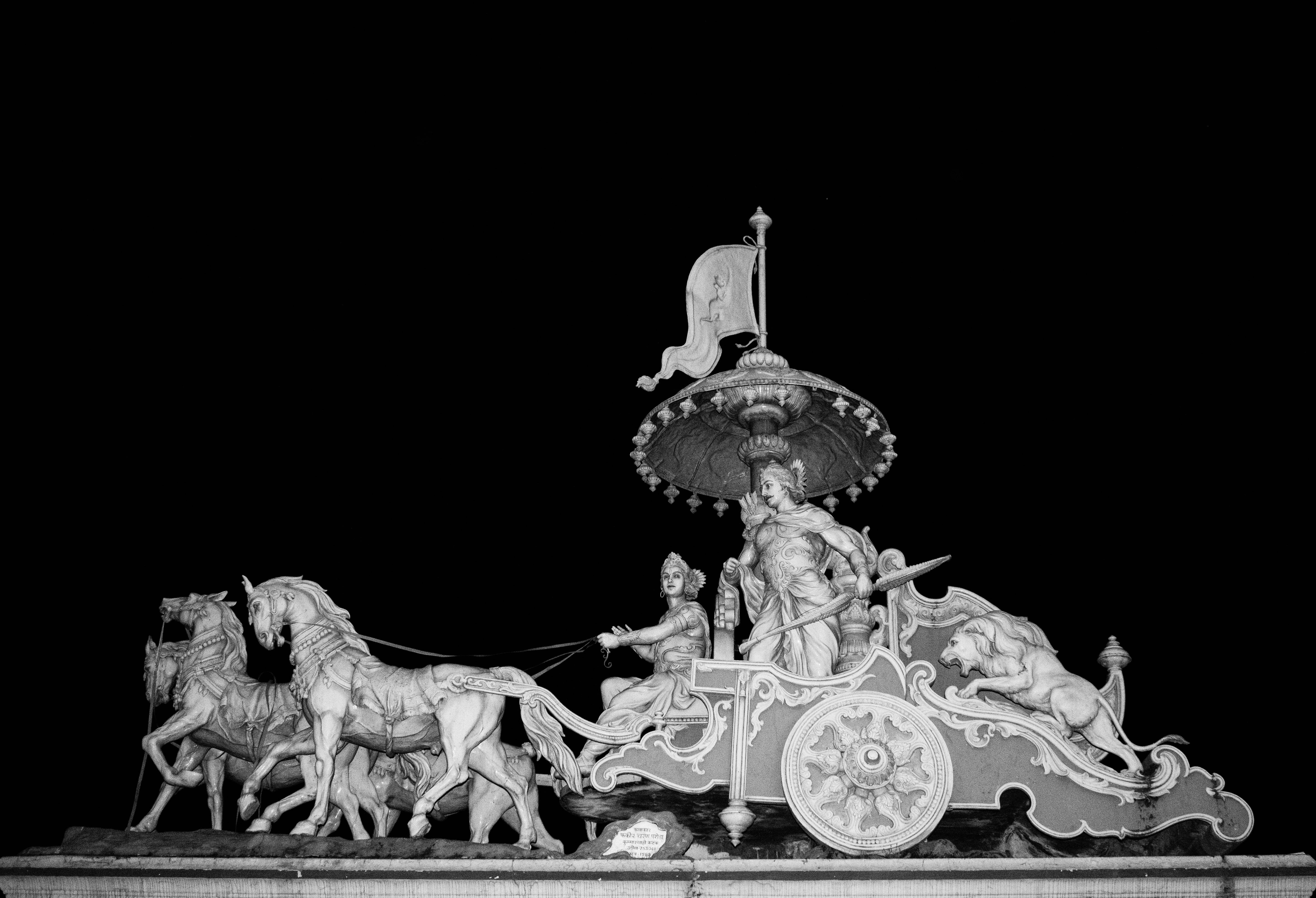 Shri Krishna 桌布 壁紙 — 一張戲劇性的黑白夜間照片，展示了一輛由四匹馬、人物和一頭獅子拉著的華麗雕刻戰車，背景是黑暗的虛空。