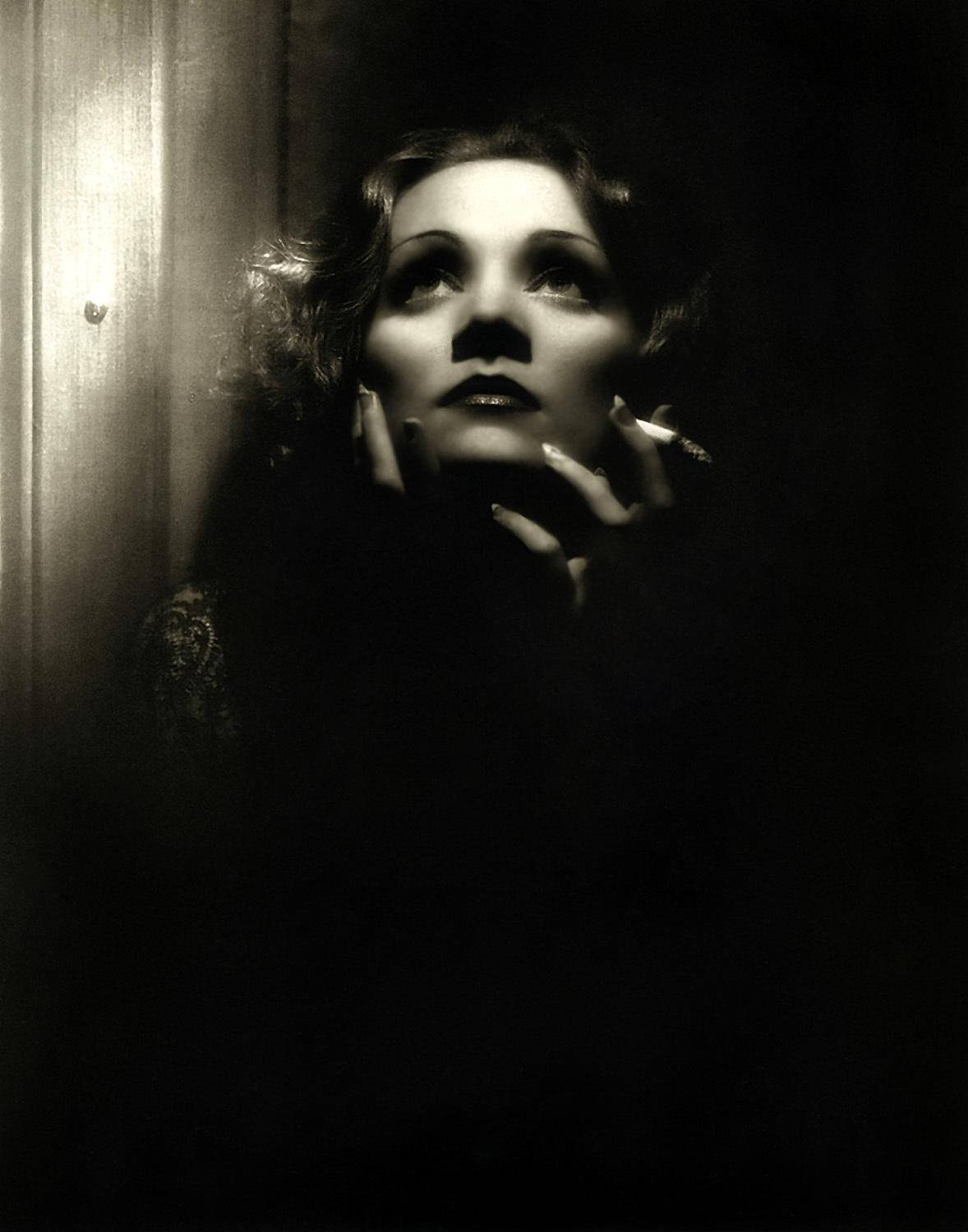 Marlene Dietrich 桌布 壁紙 — 一張戲劇性的黑白肖像，一位捲髮女性手持香菸，在昏暗的房間中向上凝視。