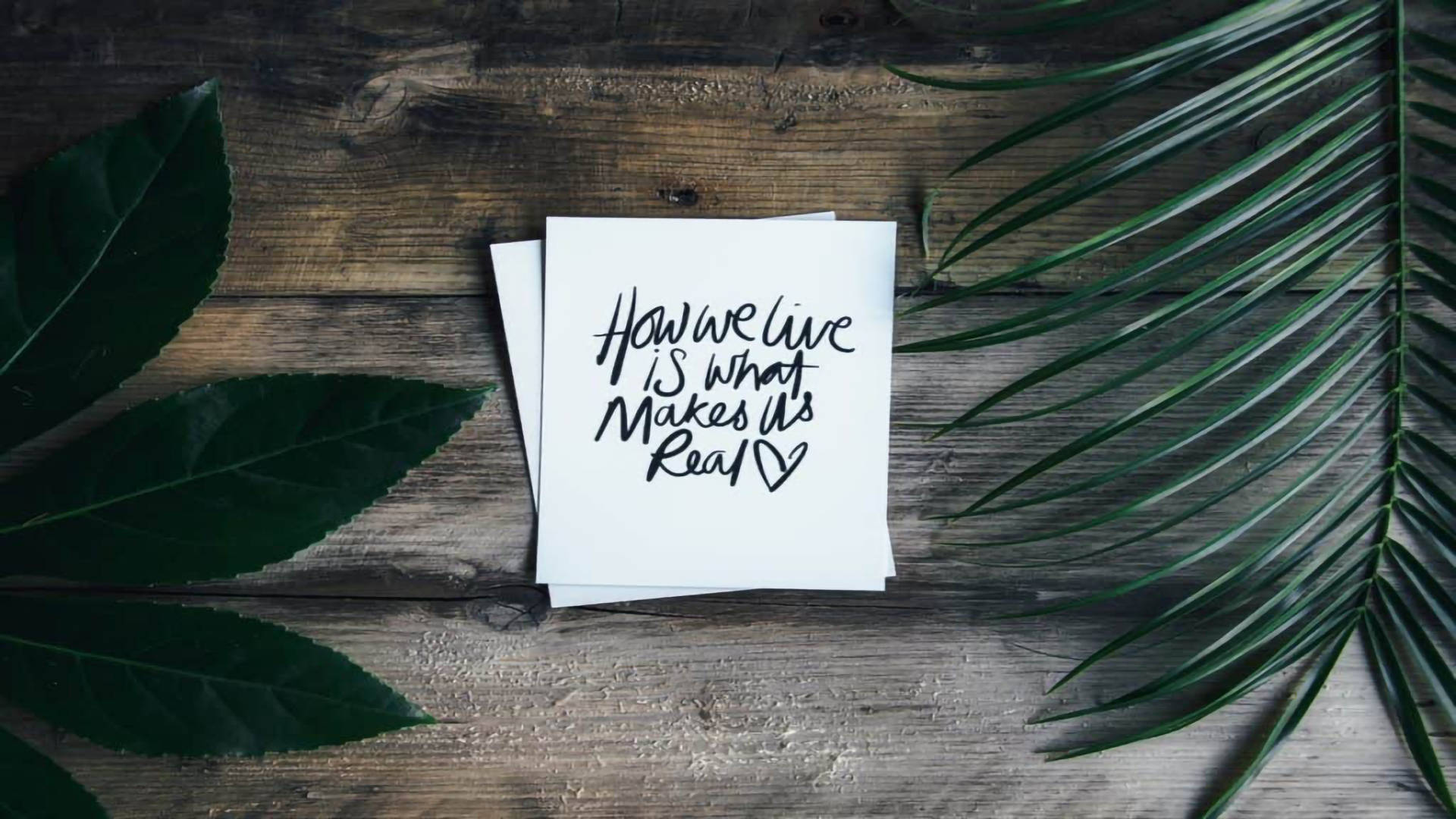Positive Quotes 桌布 壁紙 — 一張手寫便條，上面寫著「How we live is what makes it real」，放在經風化的木頭上，周圍環繞著茂盛的綠葉。