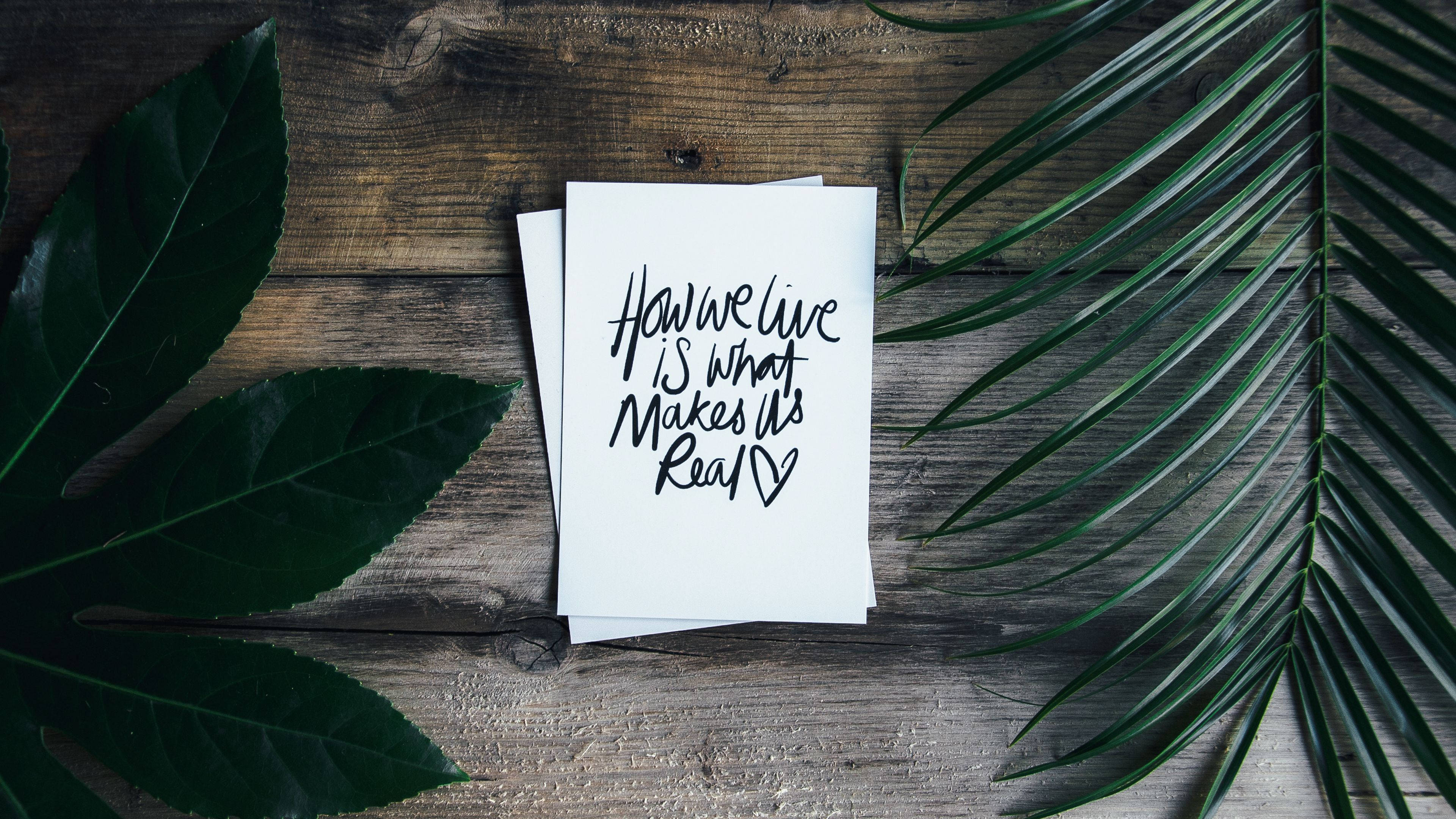 桌布 壁紙 — 一張手寫便條，上面寫著「How we live is what makes it real」，放在經風化的木頭上，周圍環繞著茂盛的綠色熱帶葉子。