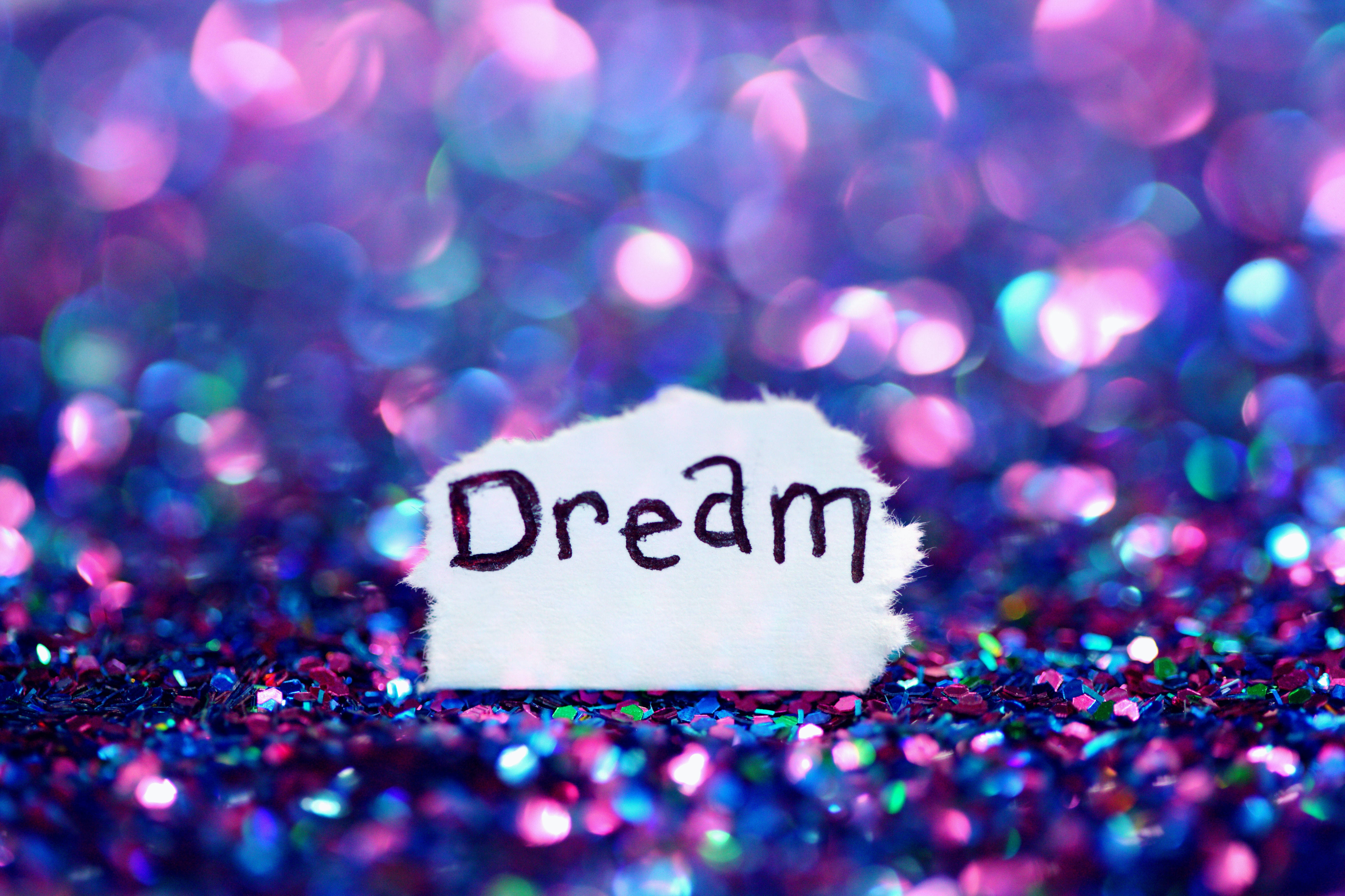 夢幻 桌布 壁紙 — 一張手寫的「Dream」字條放在閃爍的紫色和藍色亮粉床上，背景有散景燈光。