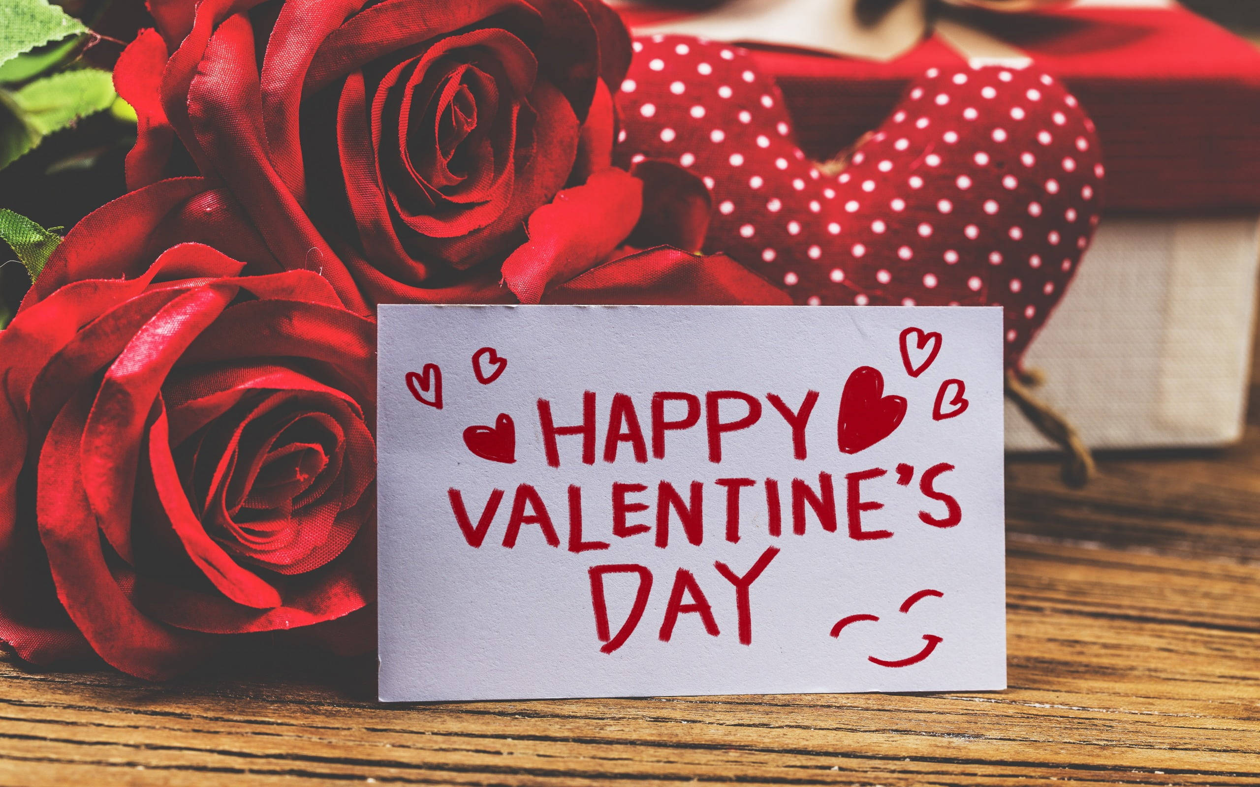 Happy Valentines Day 桌布 壁紙 — 一張手寫的心形情人節卡片放在木質表面上，旁邊有鮮紅的玫瑰和一個圓點心形。