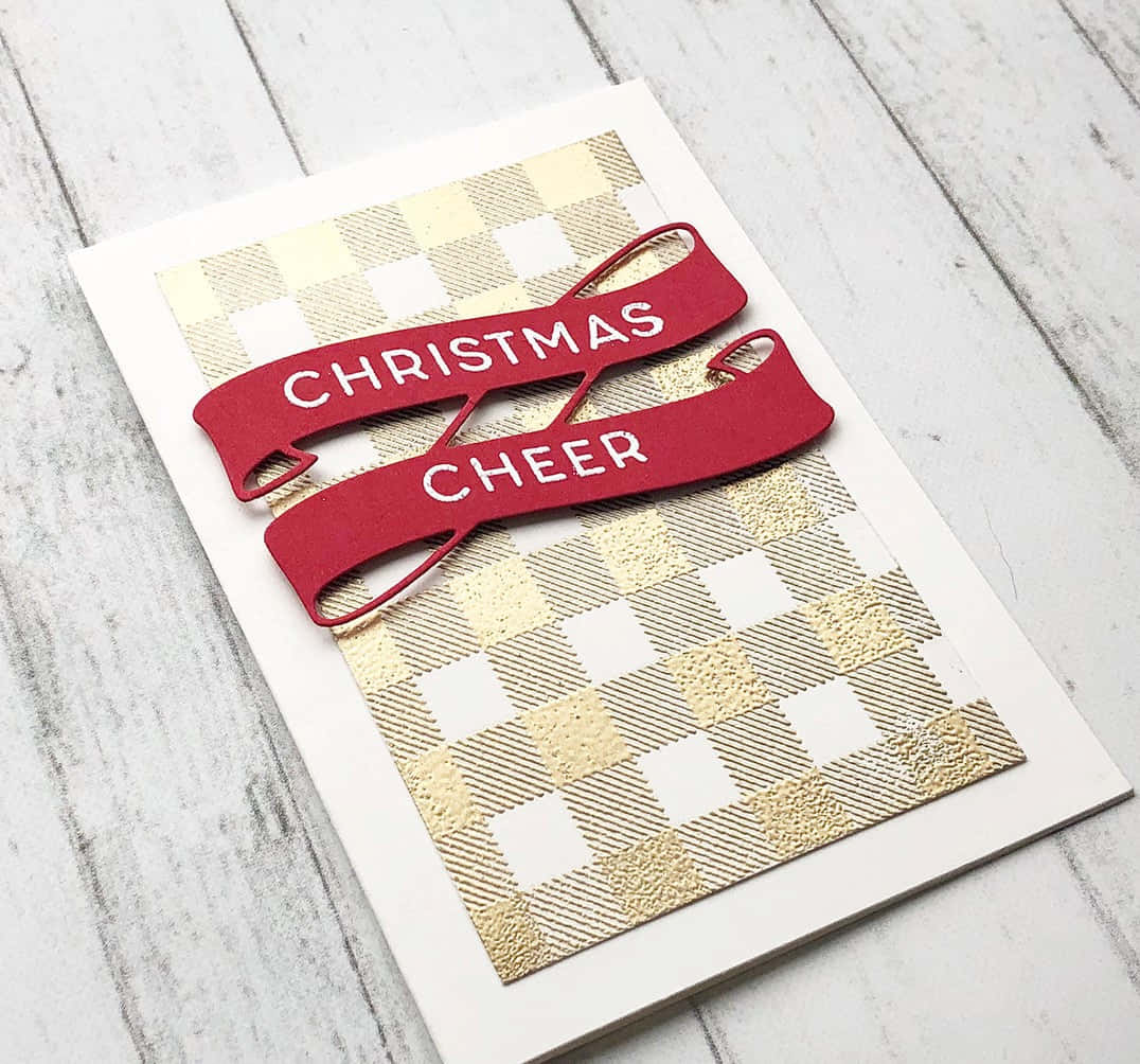Checkered 桌布 壁紙 — 一張手工聖誕卡，上面有紅色絲帶橫幅，寫著「CHRISTMAS CHEER」，位於白色木質表面上的金色和白色格子圖案之上。