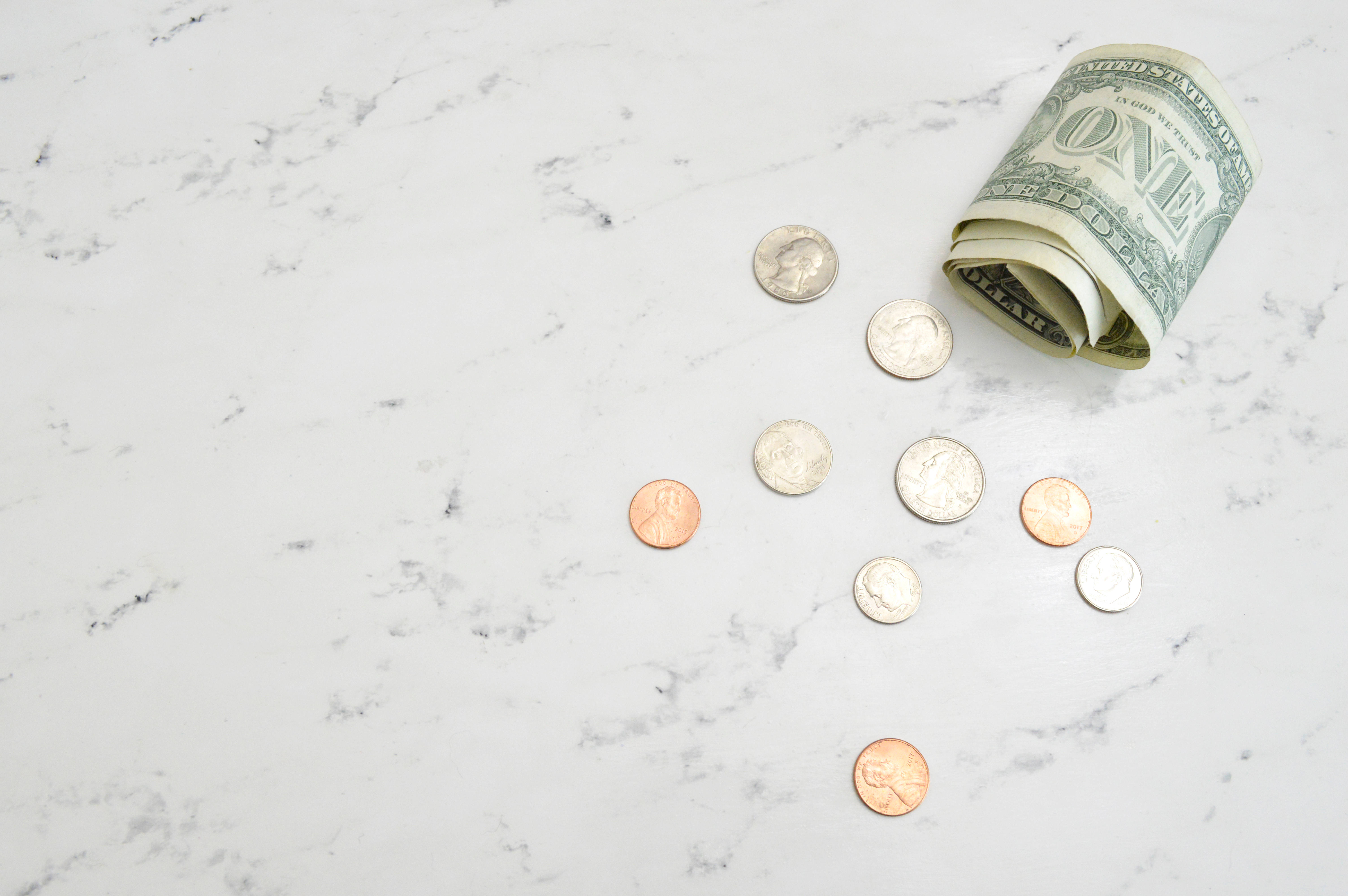 No Money 桌布 壁紙 — 一張捲起的一美元鈔票和散落的美國硬幣放在乾淨的白色大理石表面上。