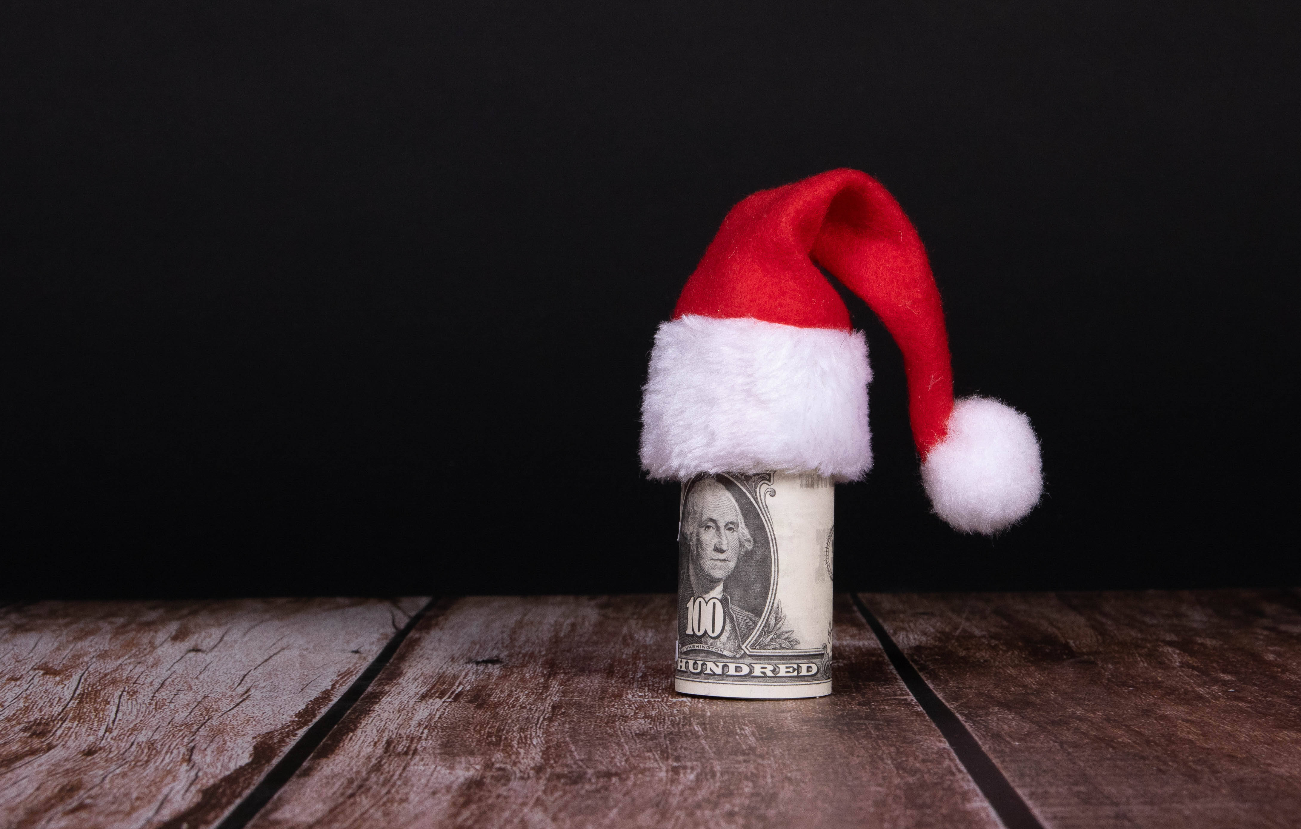 No Money 桌布 壁紙 — 一張捲起的百元美鈔戴著紅白聖誕帽，放在黑色背景前的質樸木質表面上。