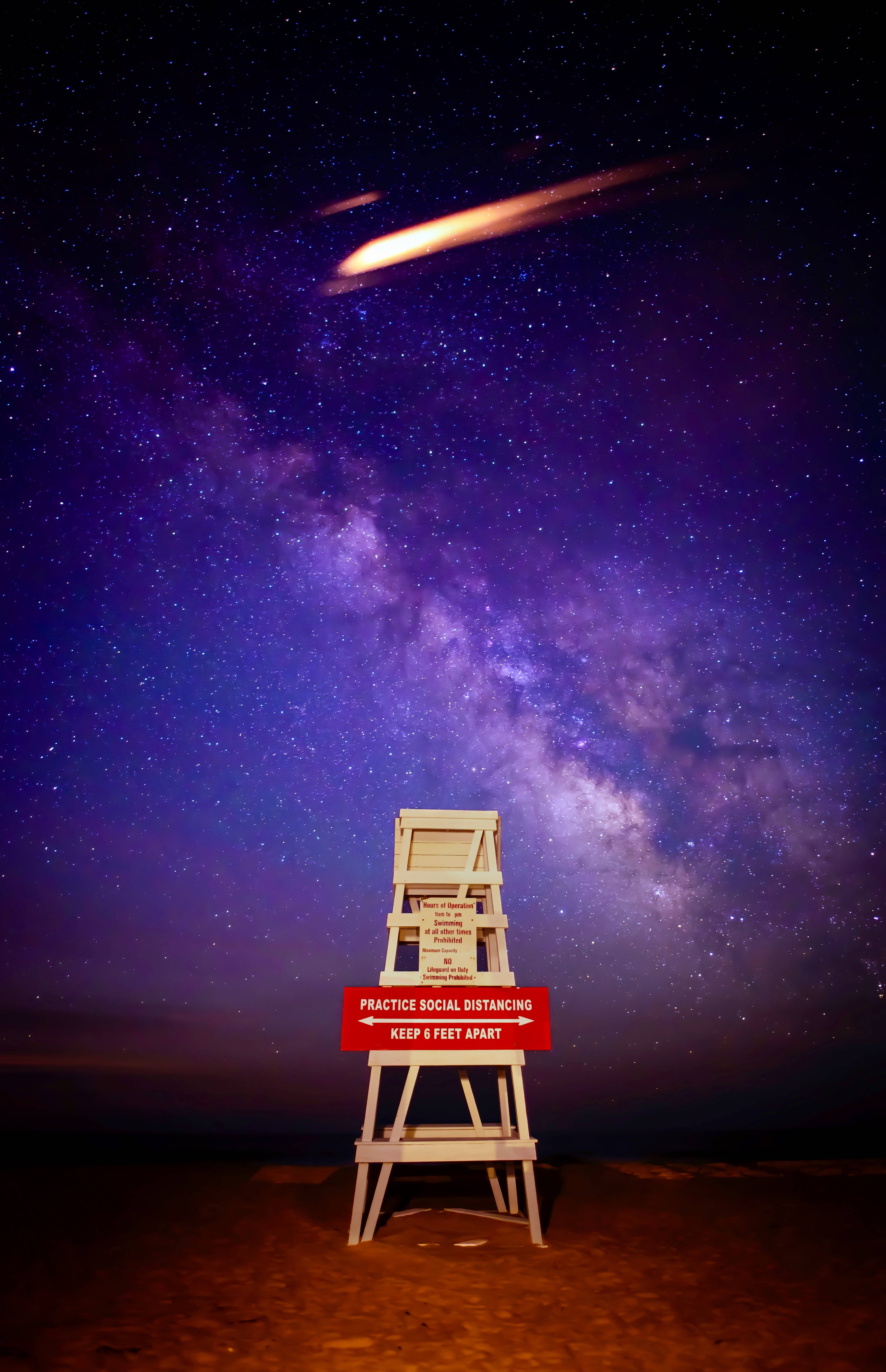 Pretty Galaxy 桌布 壁紙 — 一張救生員椅獨自矗立在黑暗的海灘上，上方是充滿活力的紫色銀河天空，劃過一顆發光的流星。