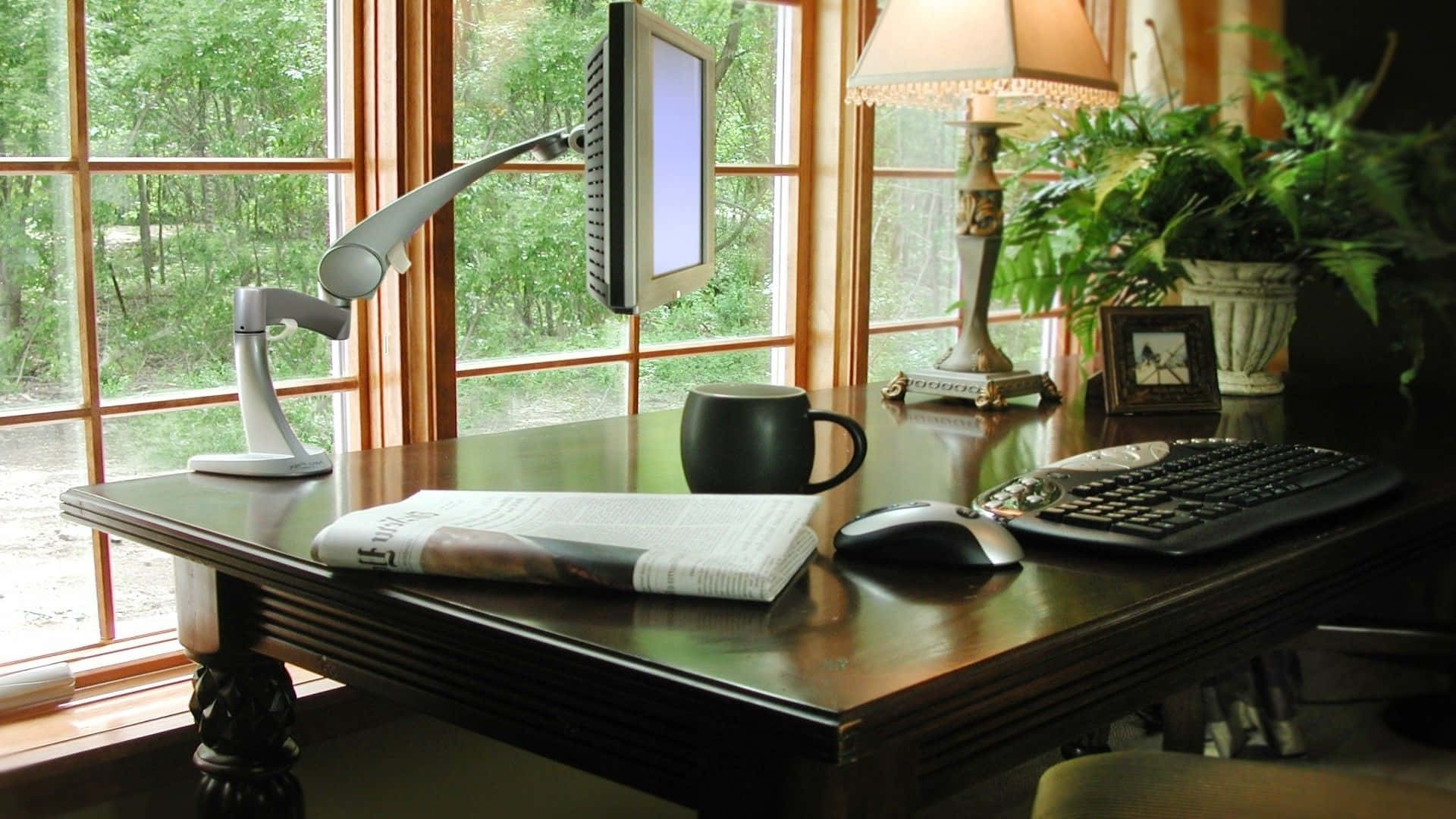 Working From Home 桌布 壁紙 — 一張木桌，上面放有顯示器、馬克杯、滑鼠和雜誌，位於可俯瞰綠色森林的窗邊。