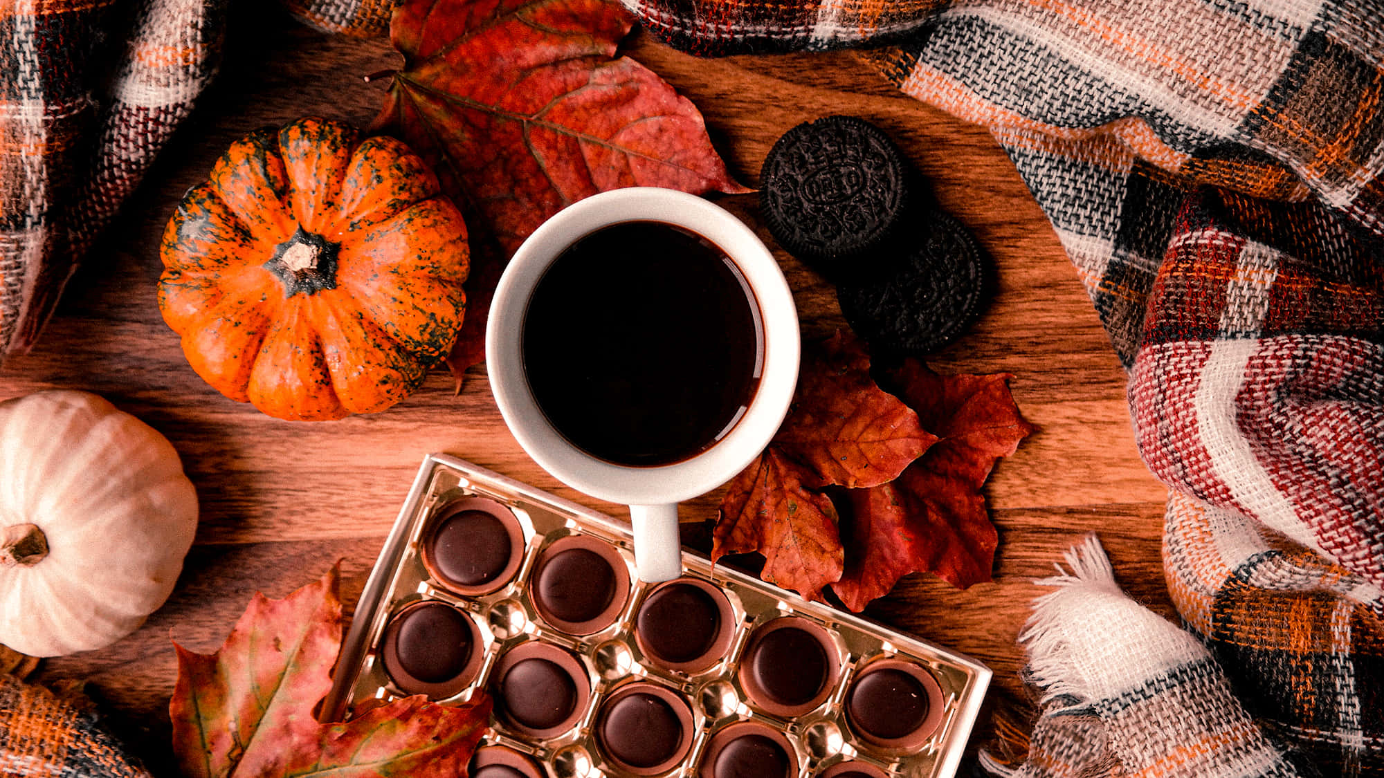 Autumn Leaves 桌布 壁紙 — 一張木桌上擺放著一杯咖啡、巧克力、南瓜和格子毯的舒適秋日場景。