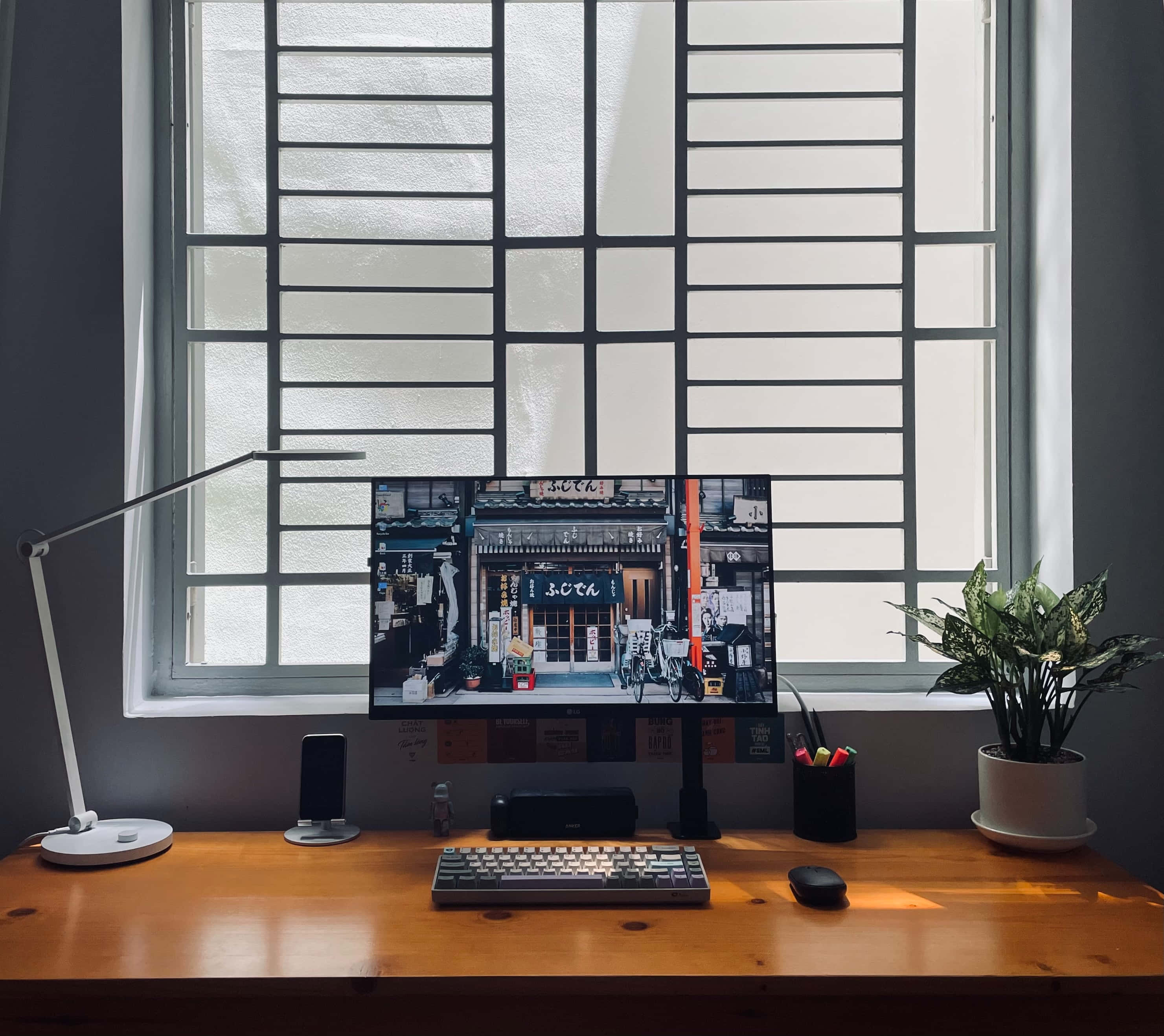 Monitor 桌布 壁紙 — 一張木製書桌，顯示器上顯示日本街景，旁邊有燈和盆栽，位於帶磨砂玻璃的大窗戶下。