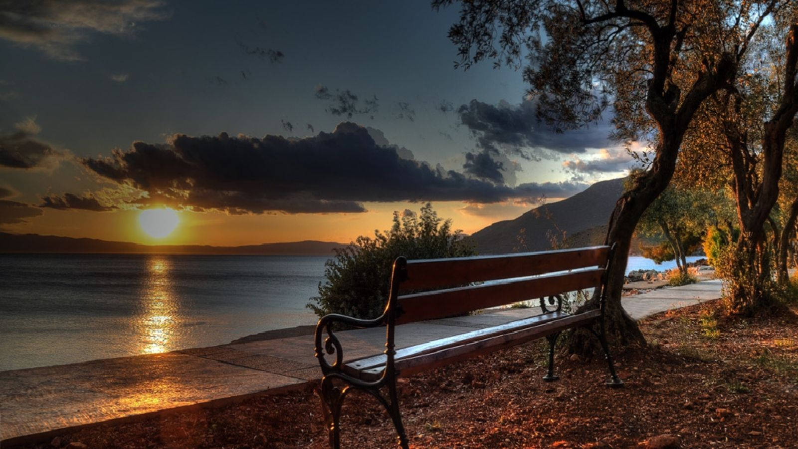 Bench 桌布 壁紙 — 一張木製長椅放在寧靜湖邊的樹下，夕陽在遠山後落下，將金色光芒灑在水面上。