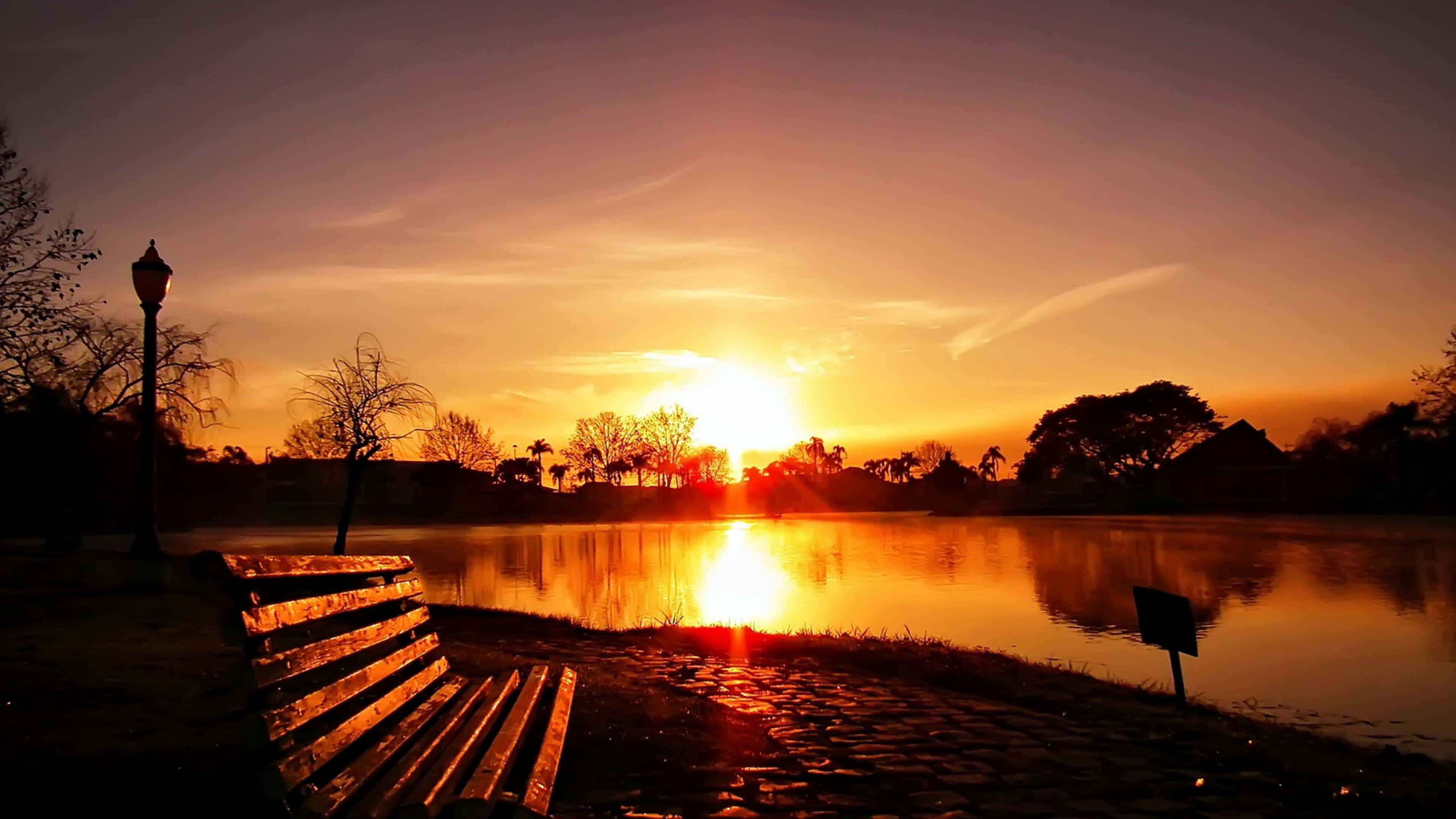 Sunset Landscape 桌布 壁紙 — 一張木製長椅面向寧靜的湖泊，夕陽西下，天空呈現火紅橙色，樹木形成剪影。