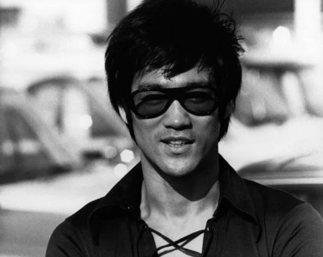 李小龍 桌布 壁紙 — 一張李小龍自信微笑的黑白肖像，戴著深色太陽眼鏡和一件交叉領口的深色襯衫。
