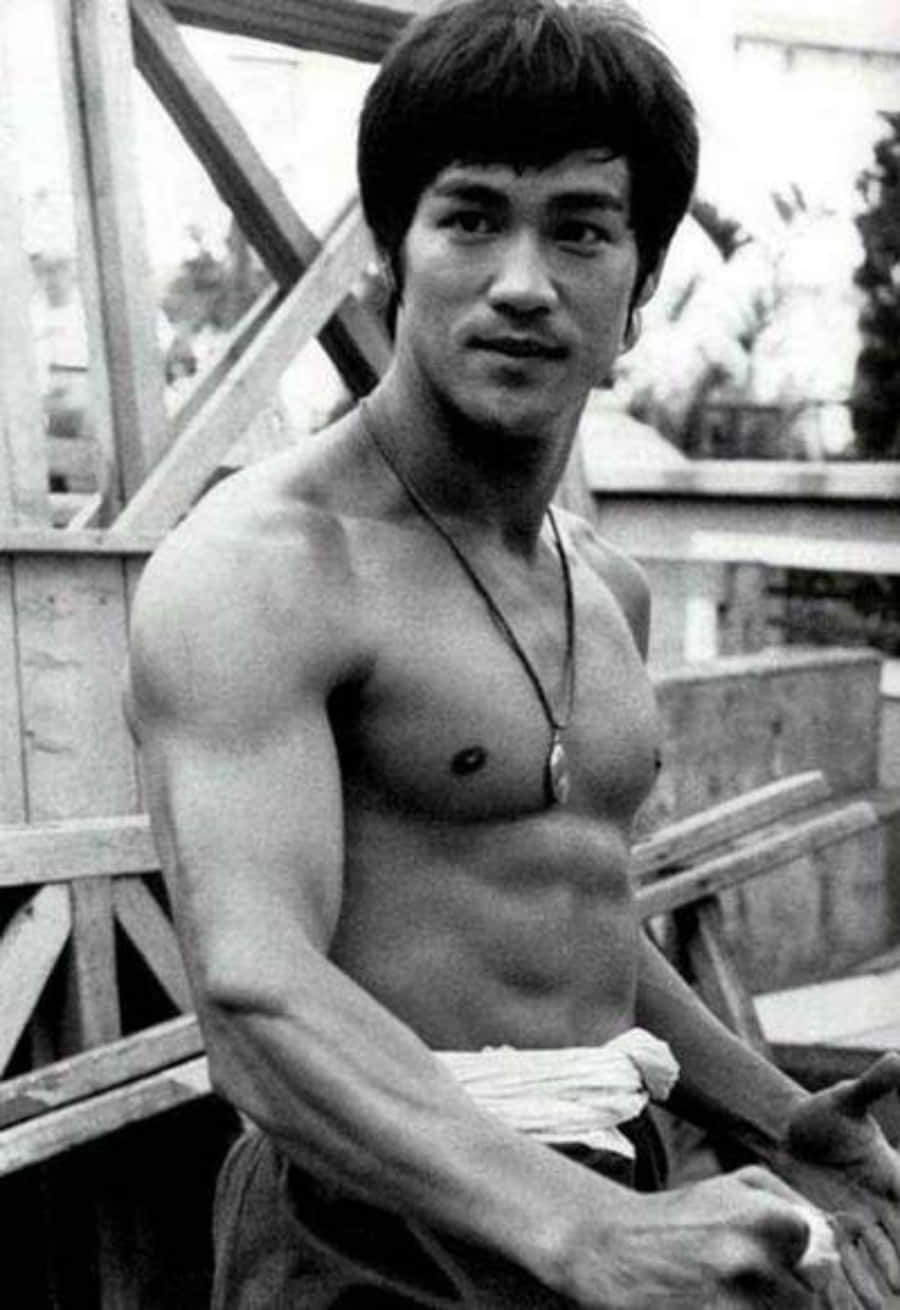 李小龍 桌布 壁紙 — 一張李小龍赤裸上身、肌肉線條分明、戴著項鍊，坐在戶外木結構附近的黑白肖像。