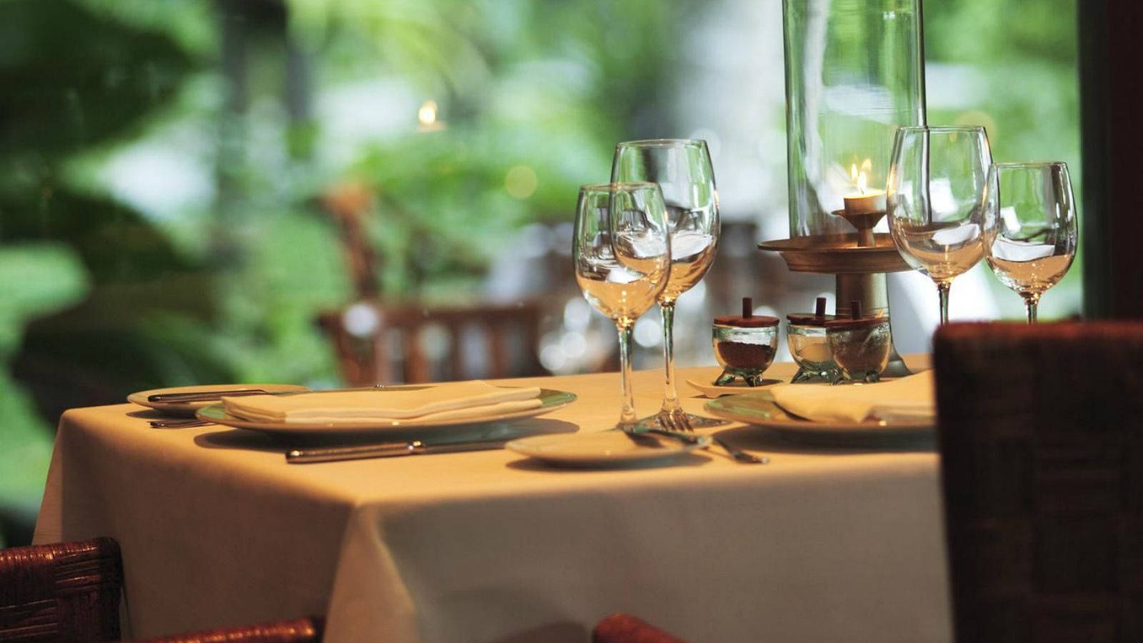 table 桌布 壁紙 — 一張柔和燈光的餐廳餐桌，擺設兩人份，有酒杯、盤子，以及一支透過室外綠植閃爍的蠟燭。
