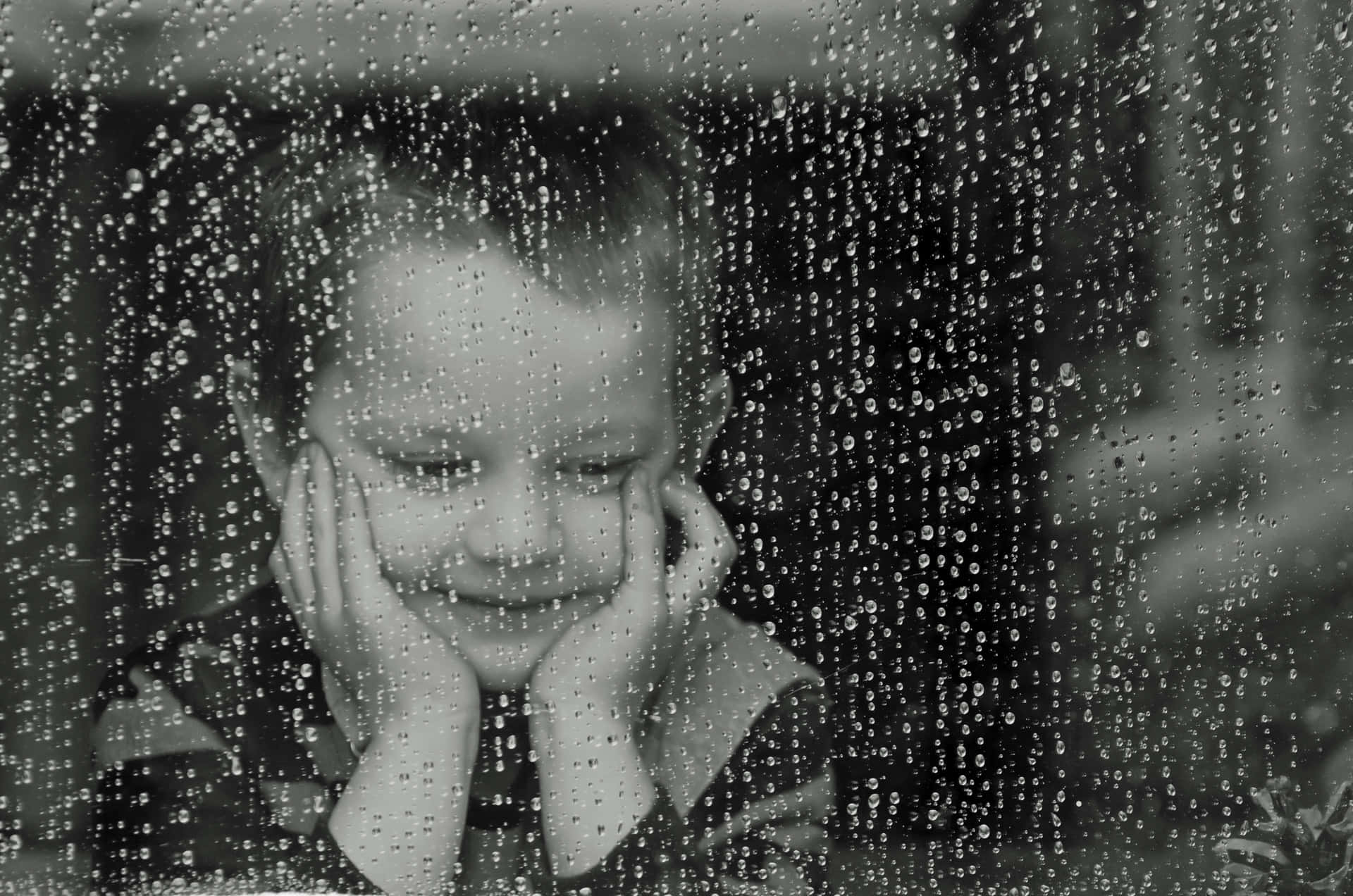 下雨 桌布 壁紙 — 一張柔和的黑白照片中，一個小孩透過沾滿雨點的窗戶溫柔地微笑。