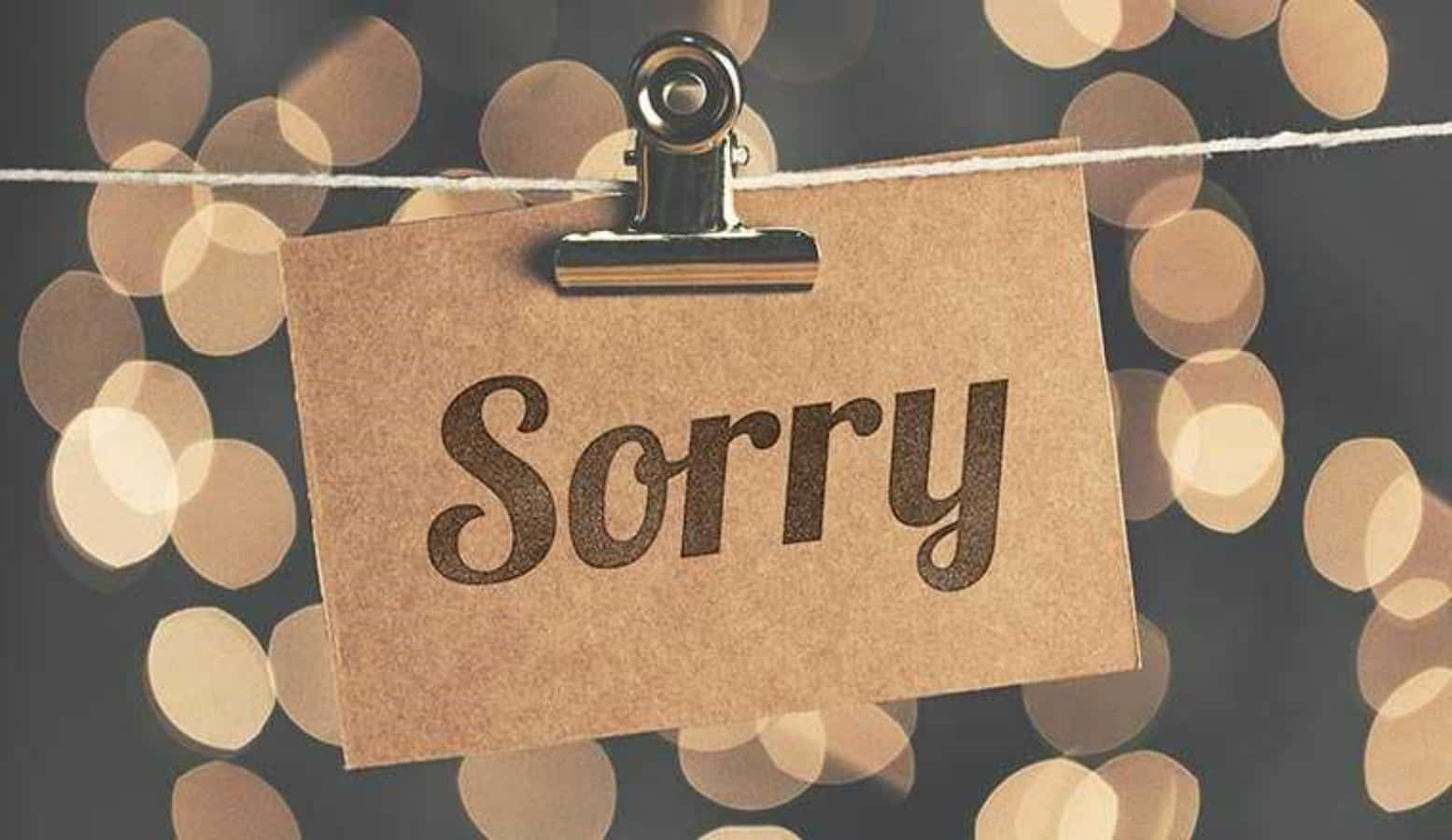 Bokeh 桌布 壁紙 — 一張棕色紙條上寫著「Sorry」，用衣夾夾在繩子上，背景是溫暖的金色散景燈光。