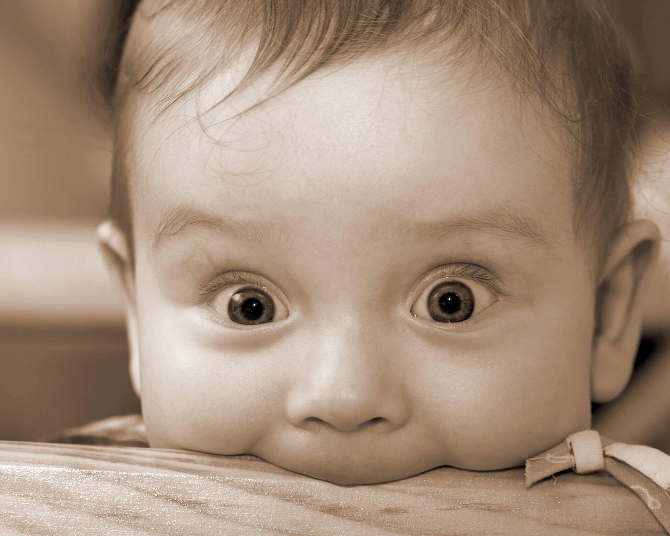 Funny Baby 桌布 壁紙 — 一張棕褐色調的特寫，嬰兒的臉龐帶著好奇的大眼睛，靠在木質表面上。
