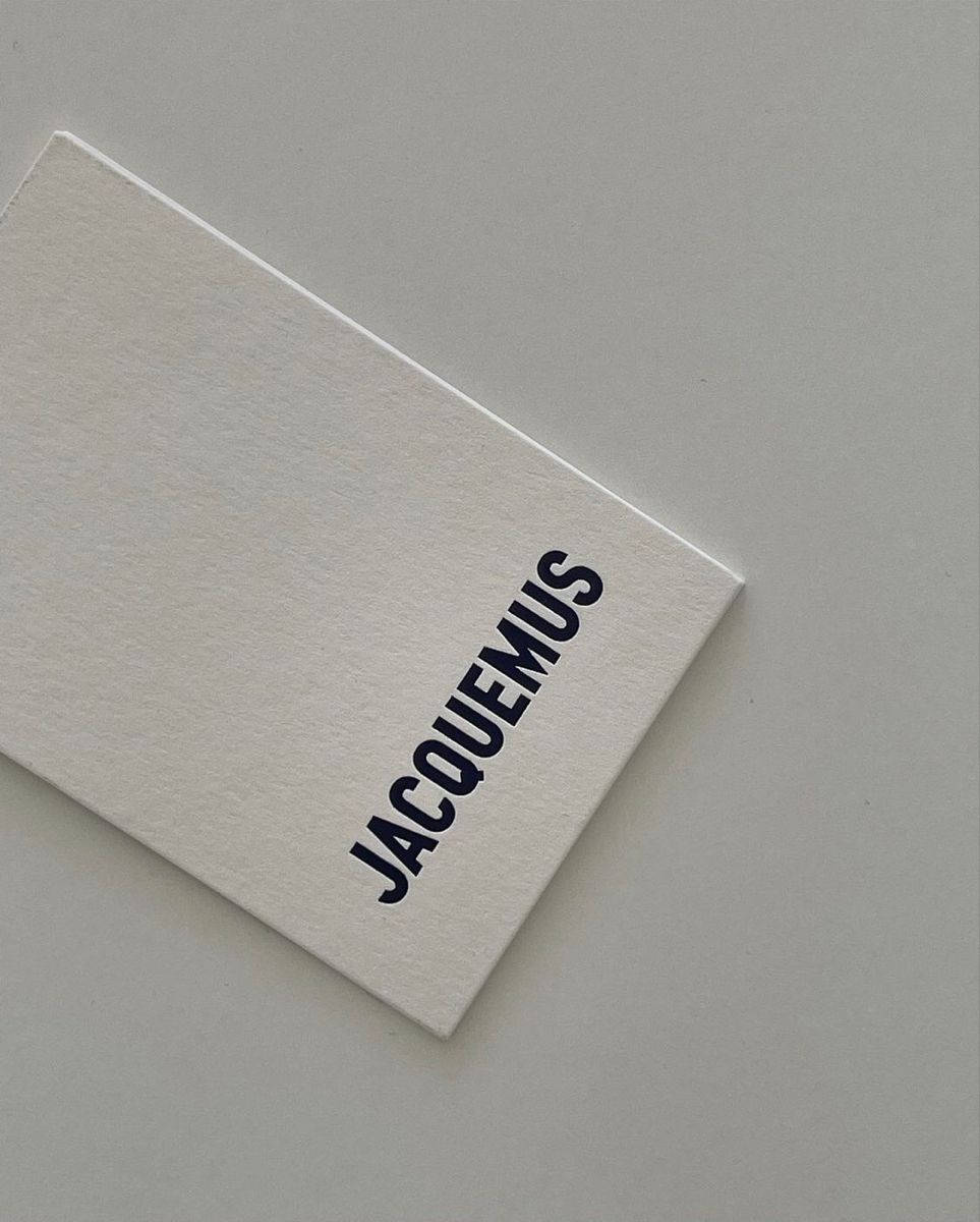 品牌 桌布 壁紙 — 一張極簡白色卡片，上面有粗體黑色「JACQUEMUS」文字，對角線放置在純灰色表面上。