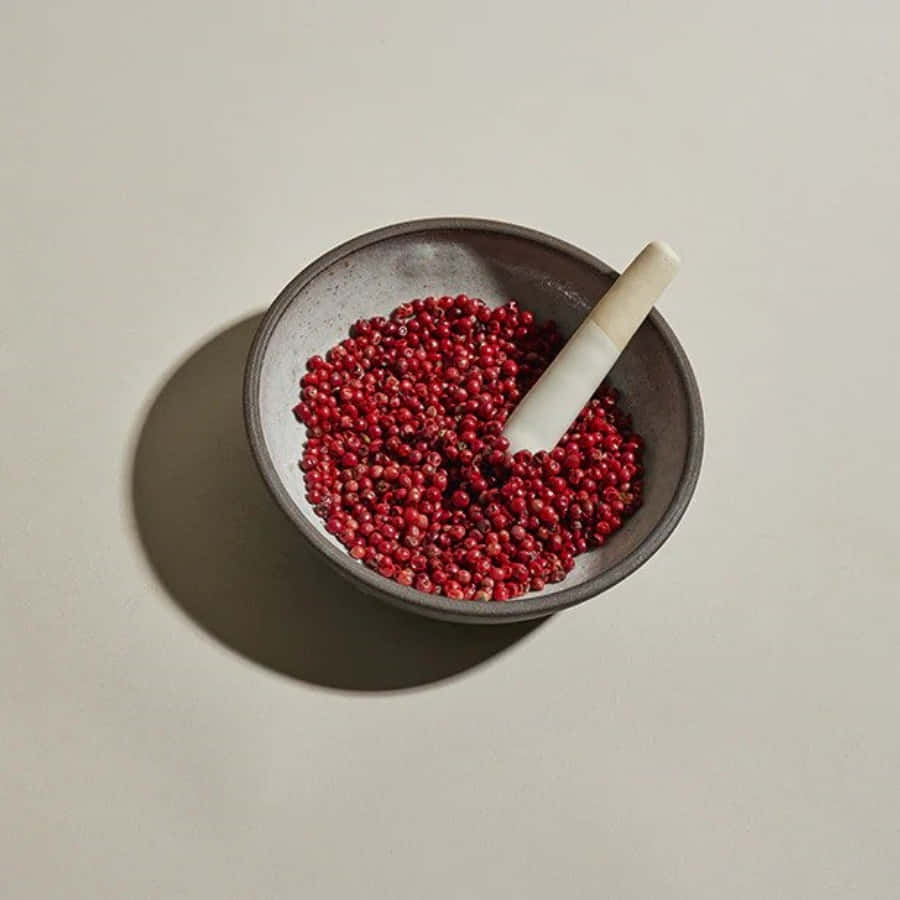 Pink Peppercorn 桌布 壁紙 — 一張極簡風格的俯拍照片，展示一個佈滿斑點的陶瓷碗，裡面裝滿了鮮紅色的胡椒粒，還有一個白色研杵，在米色中性表面上投下柔和陰影。