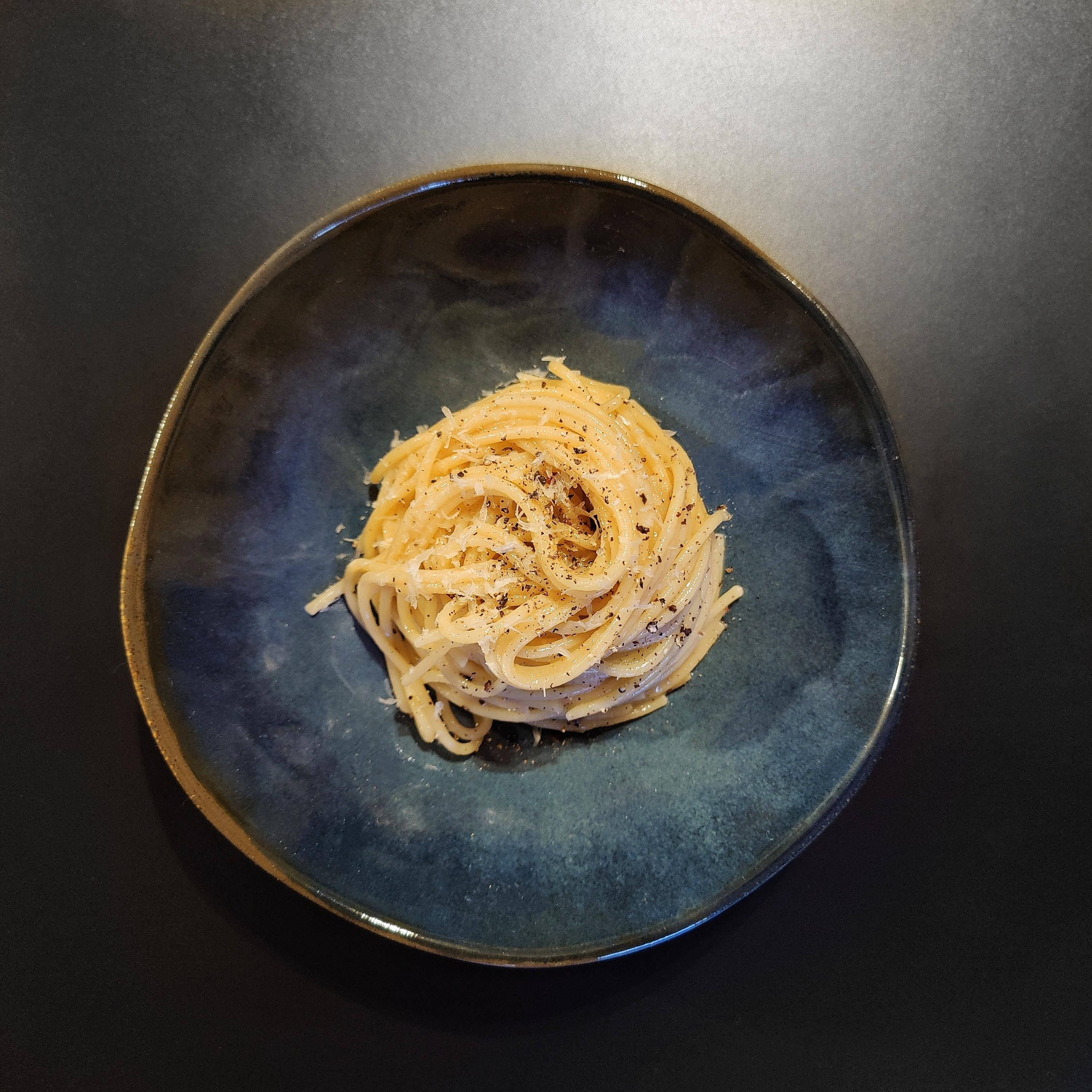 烹飪 桌布 壁紙 — 一張極簡風格的俯拍照片，展示深藍色陶瓷盤上的奶油義大利麵，飾以黑胡椒和磨碎的起司。
