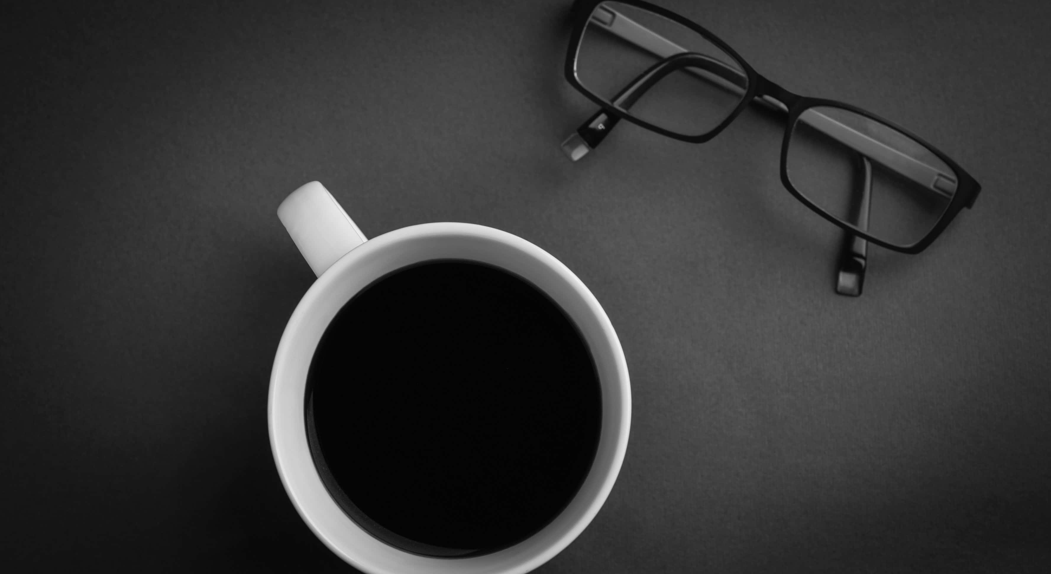 Black Coffee 桌布 壁紙 — 一張極簡黑白俯拍照片，展示一個白色咖啡杯和眼鏡放在深色紋理表面上。