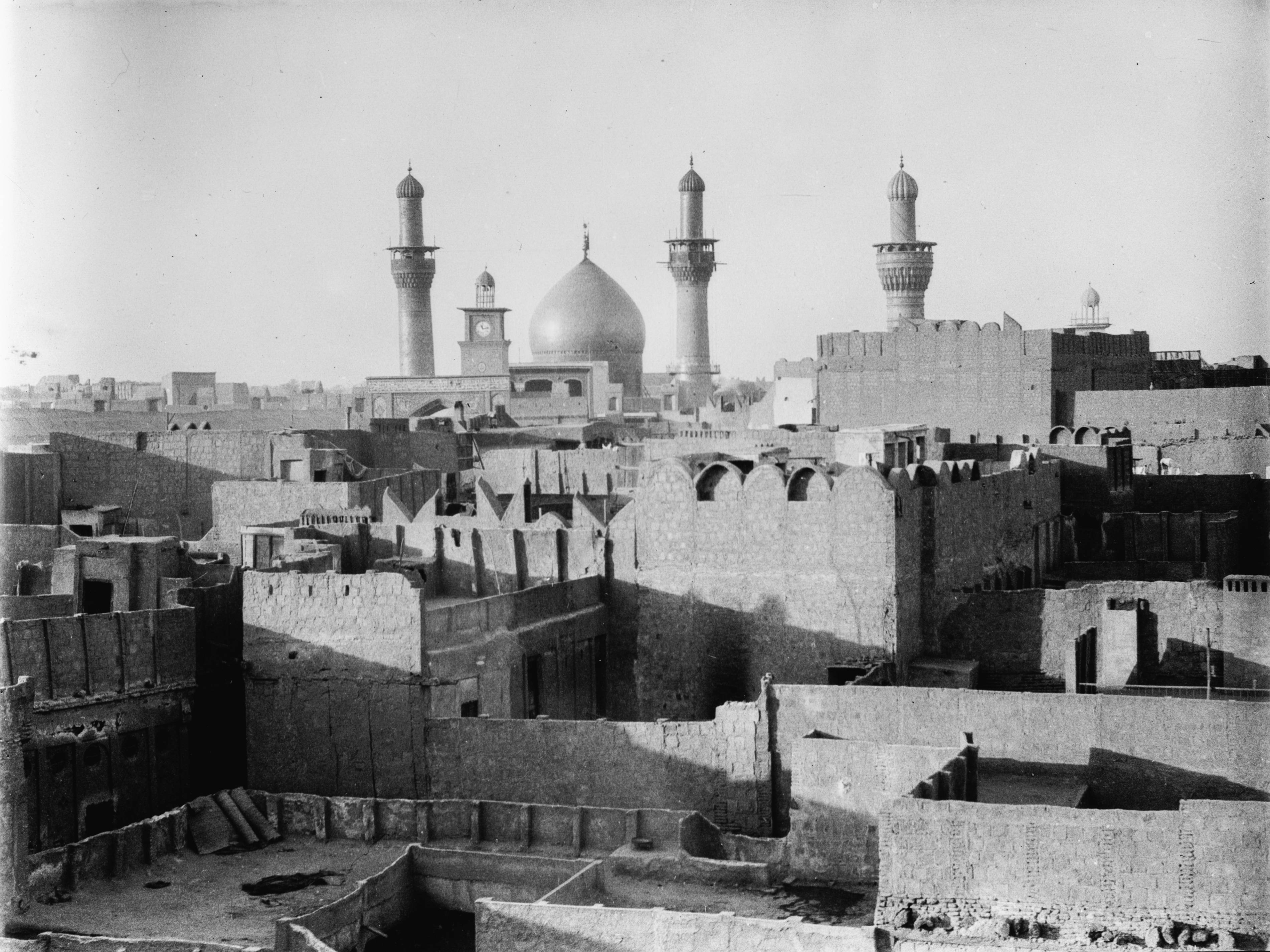 Karbala 桌布 壁紙 — 一張歷史黑白照片，顯示一座清真寺的圓頂和宣禮塔聳立於中東城市密集的舊石建築群之上。