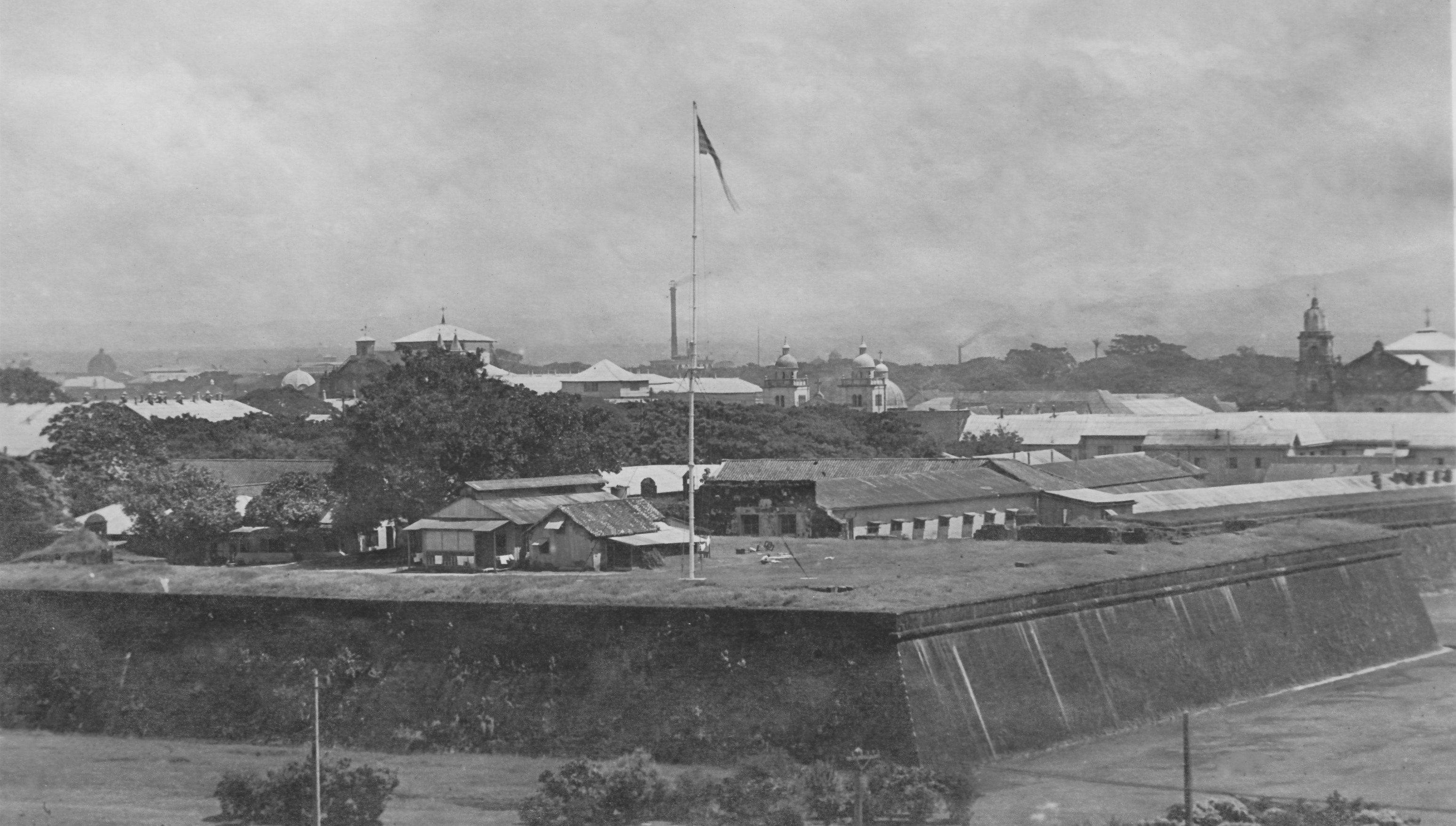 Manila 桌布 壁紙 — 一張歷史黑白照片顯示，一座設有旗杆的防禦牆俯瞰著城市景觀，其中有圓頂建築和樹木，天空多雲。