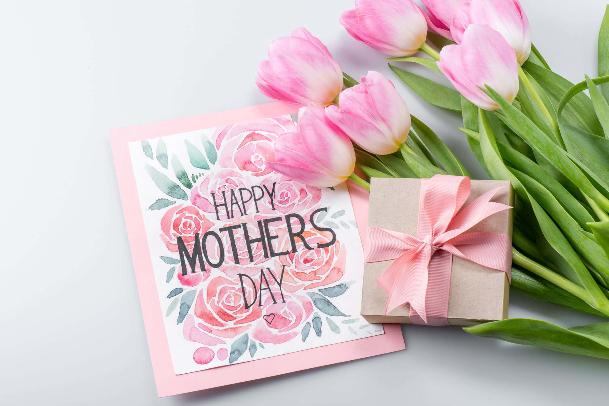 Mothers Day 桌布 壁紙 — 一張水彩母親節卡，上面有粉紅玫瑰，旁邊是粉紅鬱金香和一個用粉紅絲帶綁著的禮物盒，放在白色表面上。