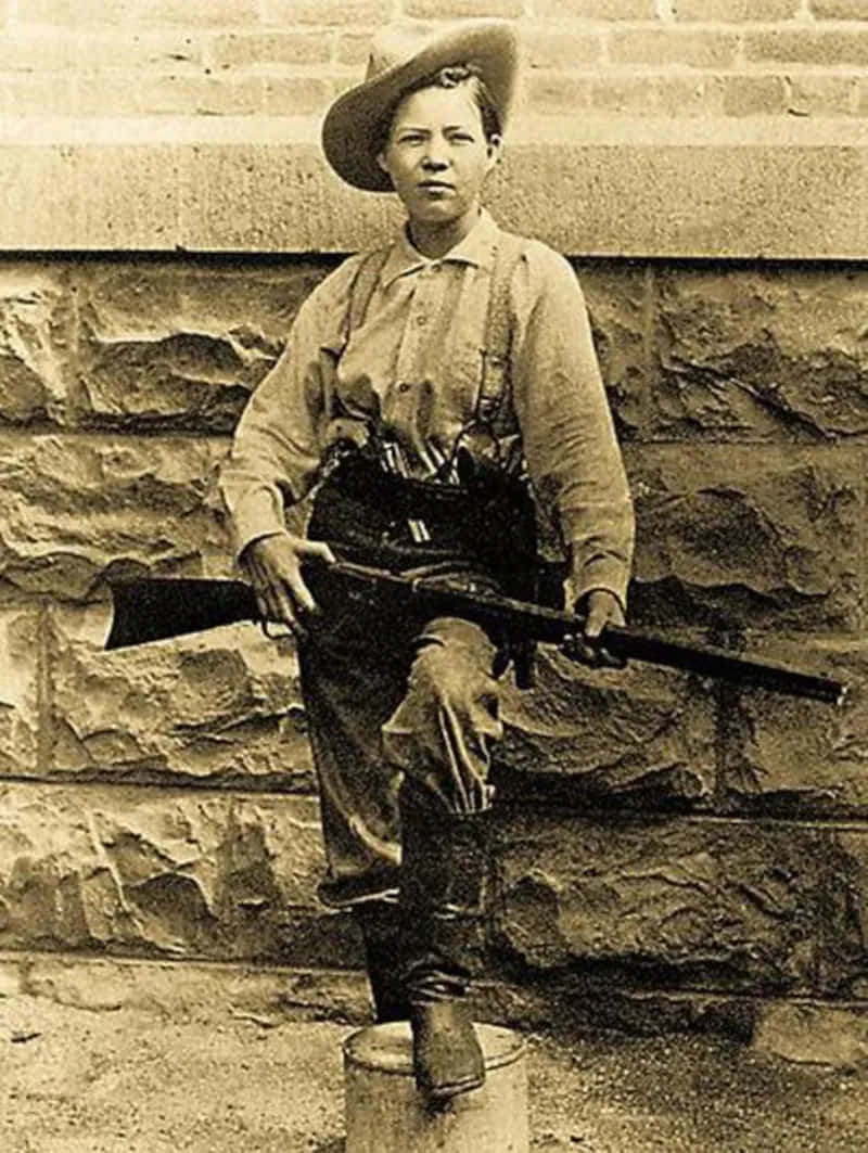 Old Western 桌布 壁紙 — 一張泛黃色調的肖像，描繪一位身穿20世紀初軍服、手持步槍、坐在石牆前的木桶上的女性。