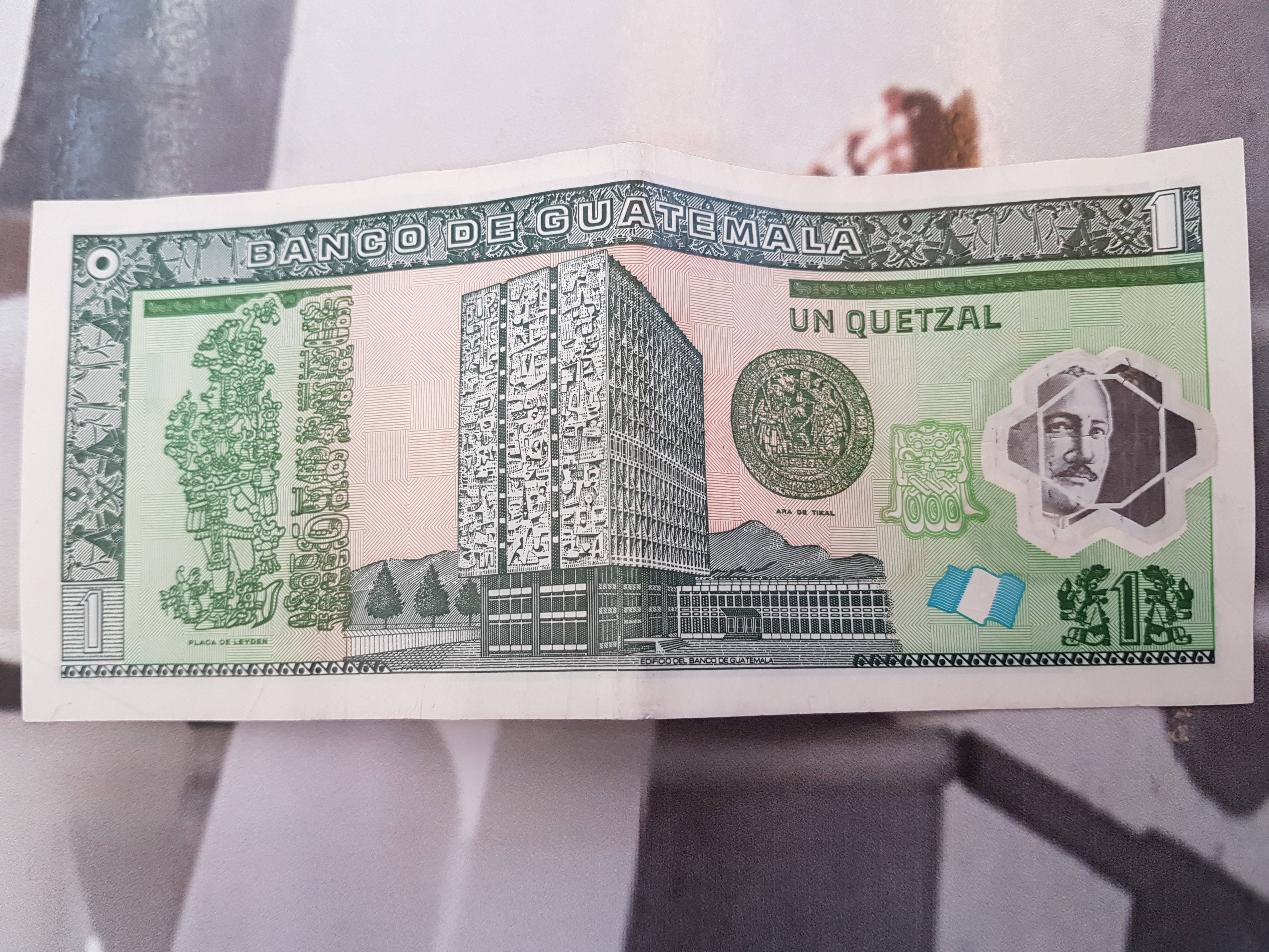 Guatemala 桌布 壁紙 — 一張清晰的瓜地馬拉一格查爾紙幣，上面有現代建築和國徽，放置在模糊的抽象背景上。