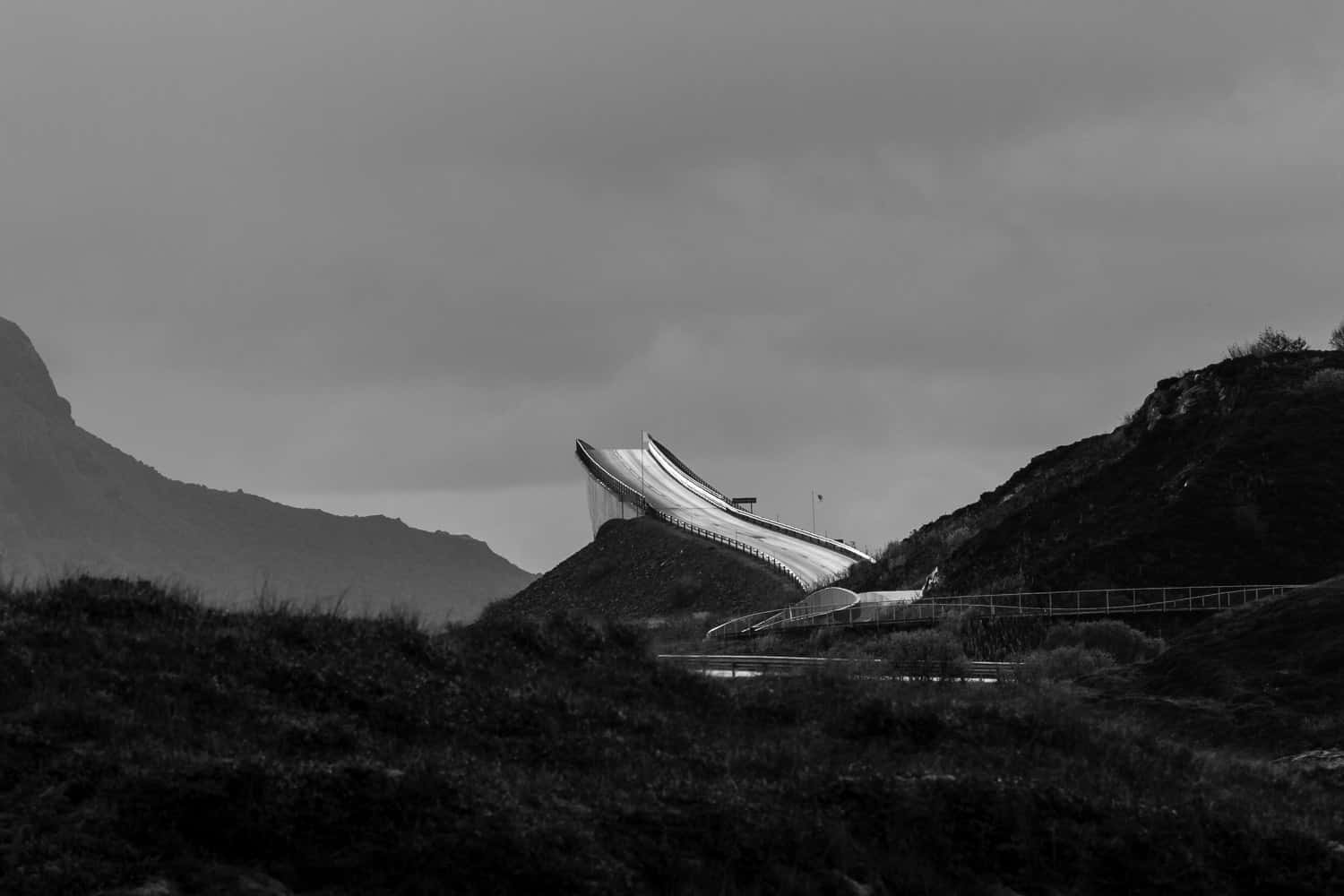 風景 桌布 壁紙 — 一張清晰的黑白照片，展示現代滑雪跳台坐落於崎嶇山丘之間，天空陰沉密佈。
