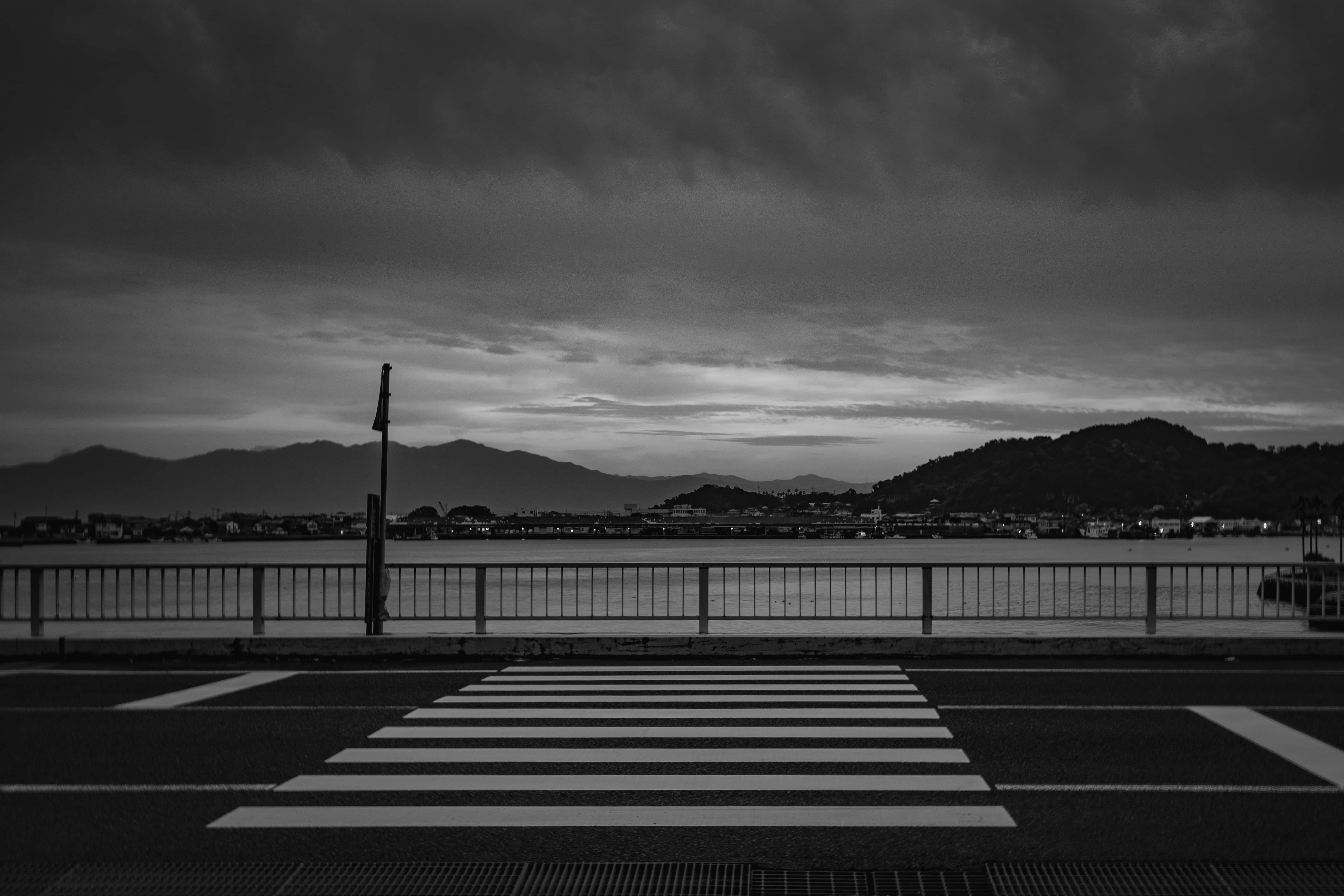 Aesthetic Black And White 桌布 壁紙 — 一張清晰的黑白照片，拍攝了一條斑馬線，通往遠處有山丘的海濱，天空烏雲密佈。