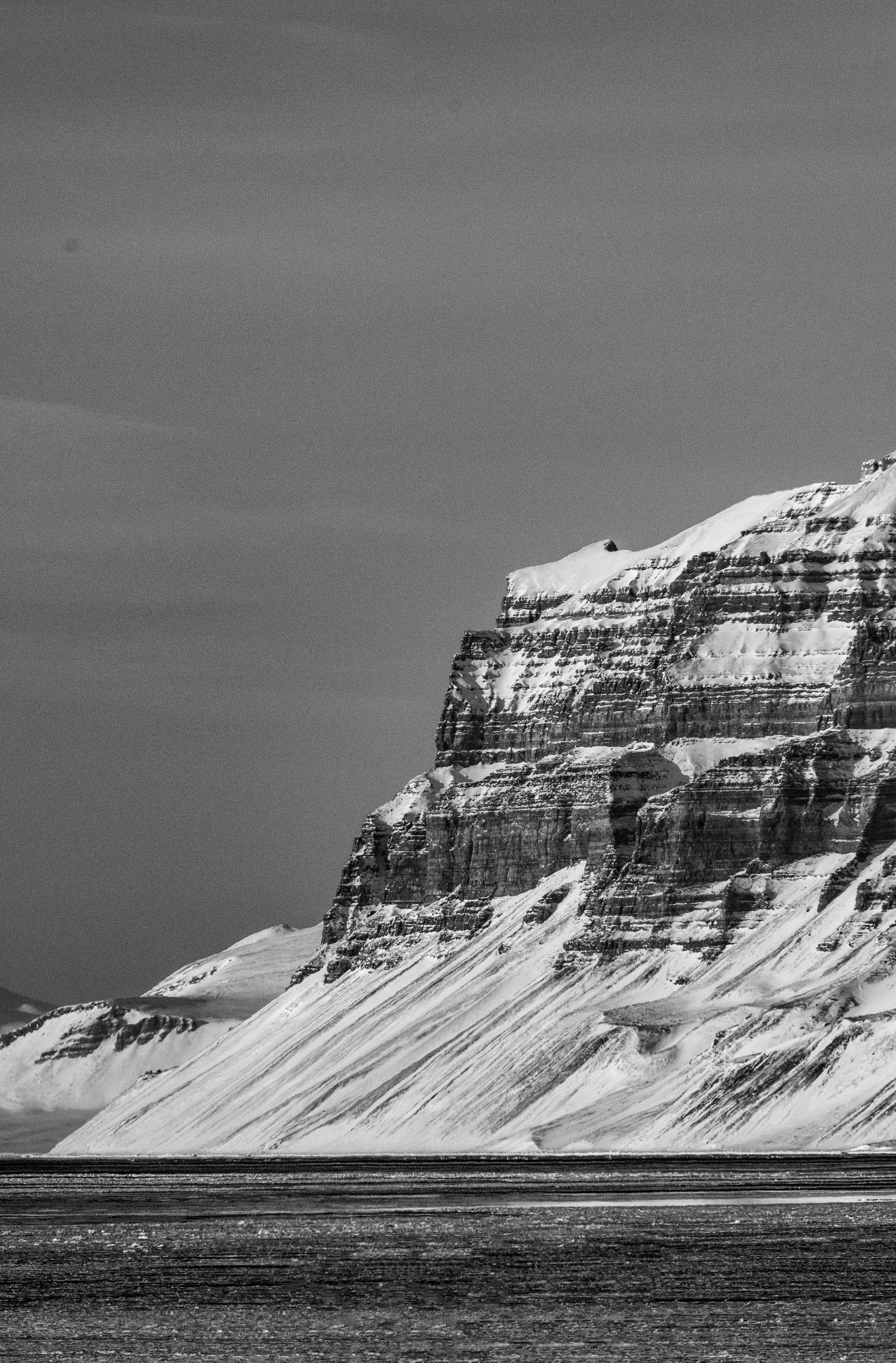 Svalbard 桌布 壁紙 — 一張清晰的黑白照片，拍攝了在陰天下一片平靜海面上聳立的積雪懸崖面。