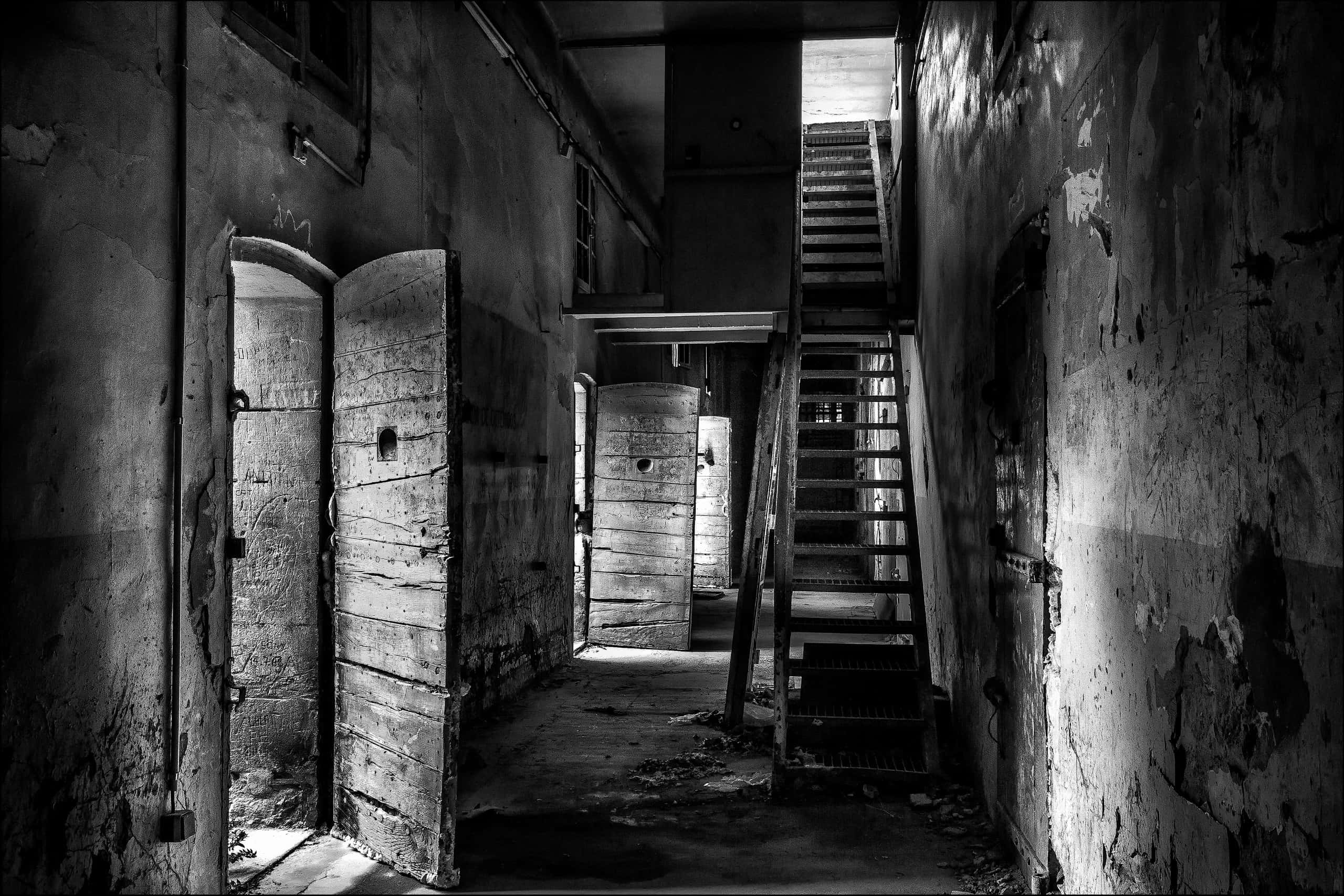 Prison Cell 桌布 壁紙 — 一張清晰的黑白照片，拍攝了腐朽的監獄走廊，有生鏽的金屬門和通往昏暗光線的樓梯。
