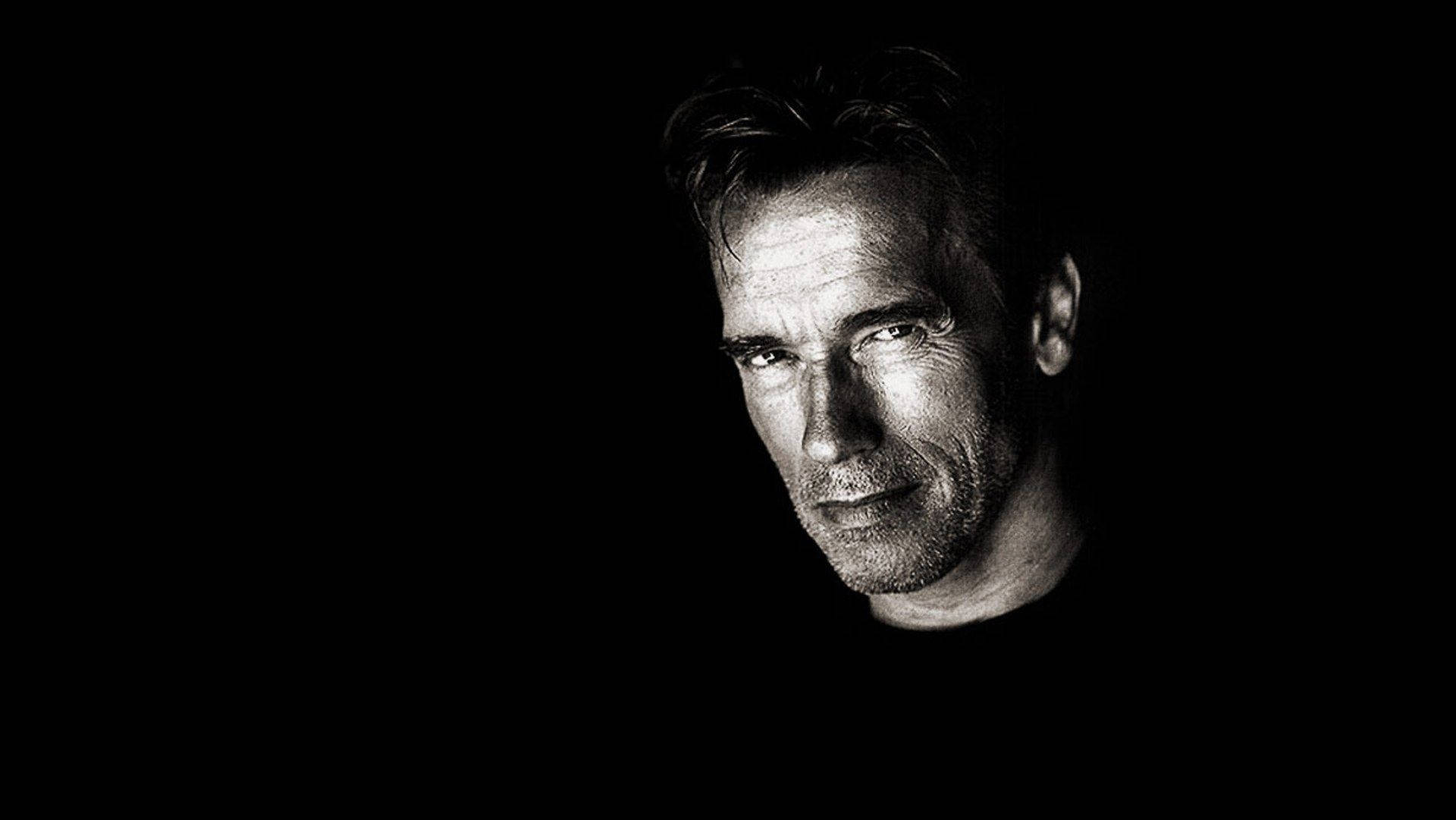 Arnold 桌布 壁紙 — 一張清晰的黑白肖像，捕捉到一個男人的臉從黑暗中浮現，眼神銳利，表情嚴肅。